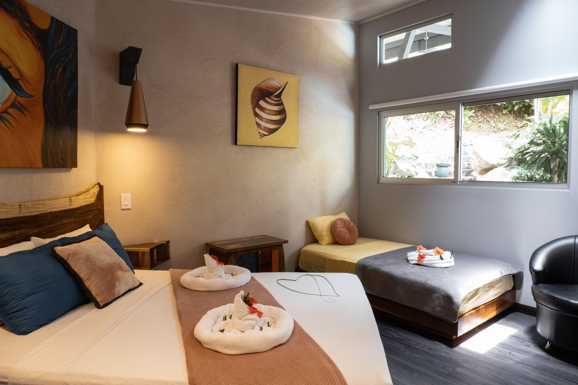 A hotel room with a queen bed and a smaller single bed, neutral walls, natural light, and modern wooden accents.