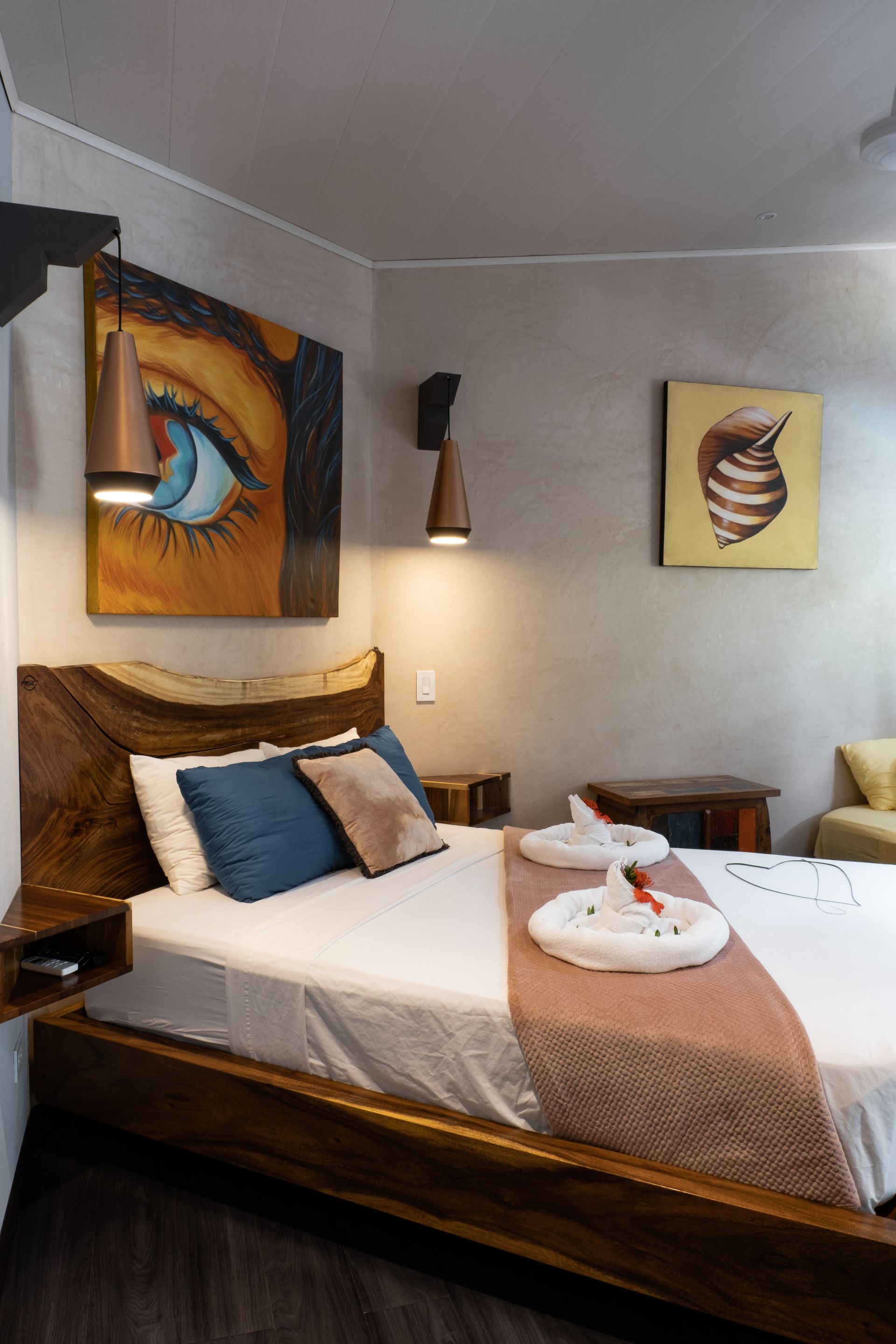 A modern hotel room with a live-edge wood bed frame, an abstract eye painting, and folded towel swans on the bed.