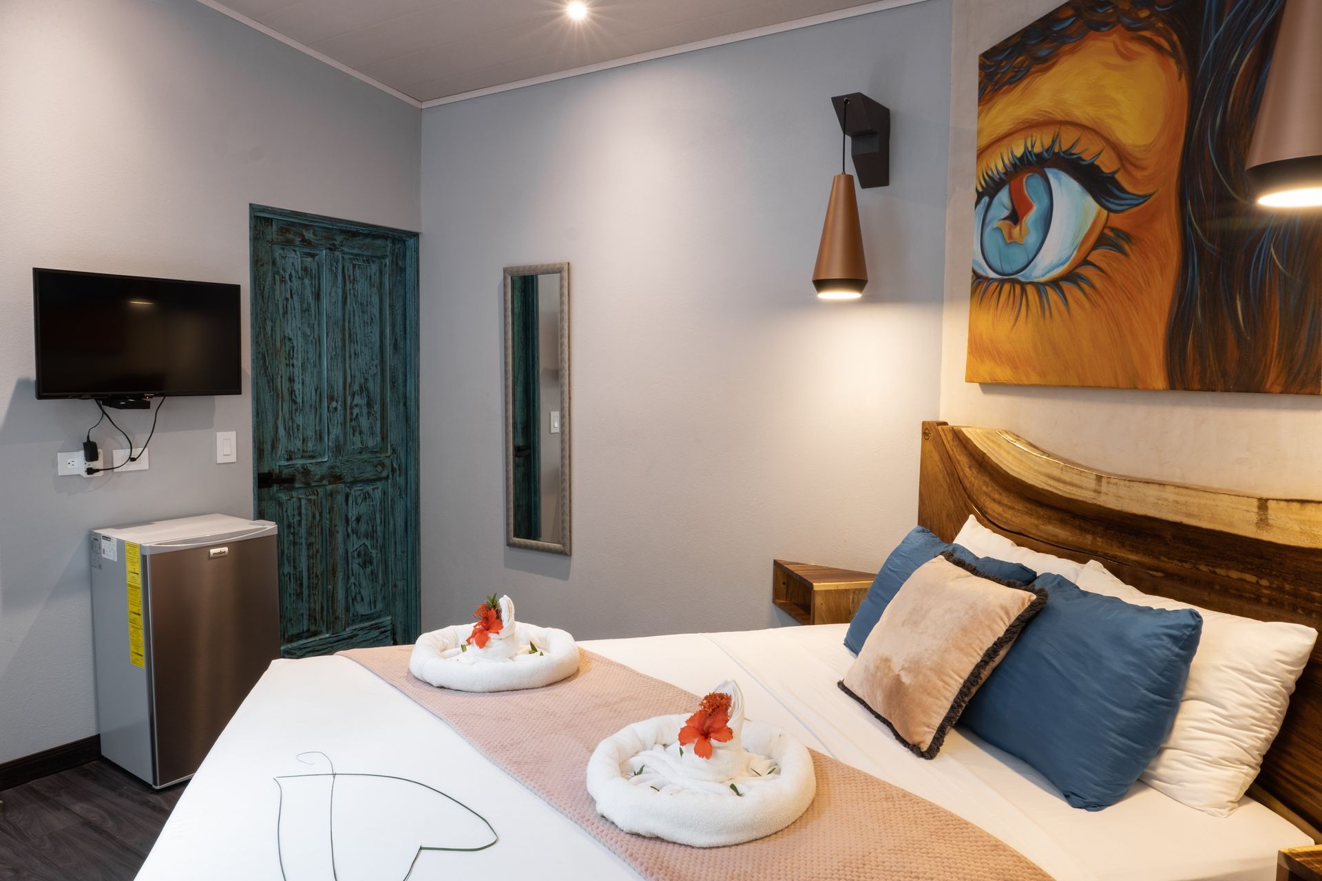 A modern hotel room with a bed featuring rolled towels, a wall-mounted TV, a mini-fridge, and a large eye painting.