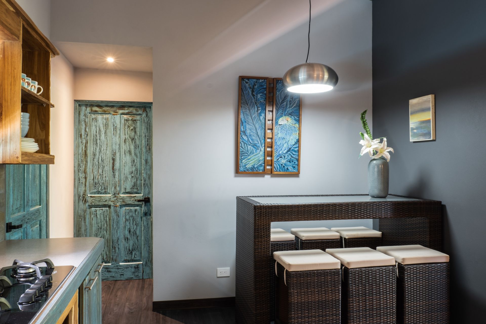 A kitchen area featuring a dark wicker table with cushioned stools, a distressed teal wooden door, and a pendant light.