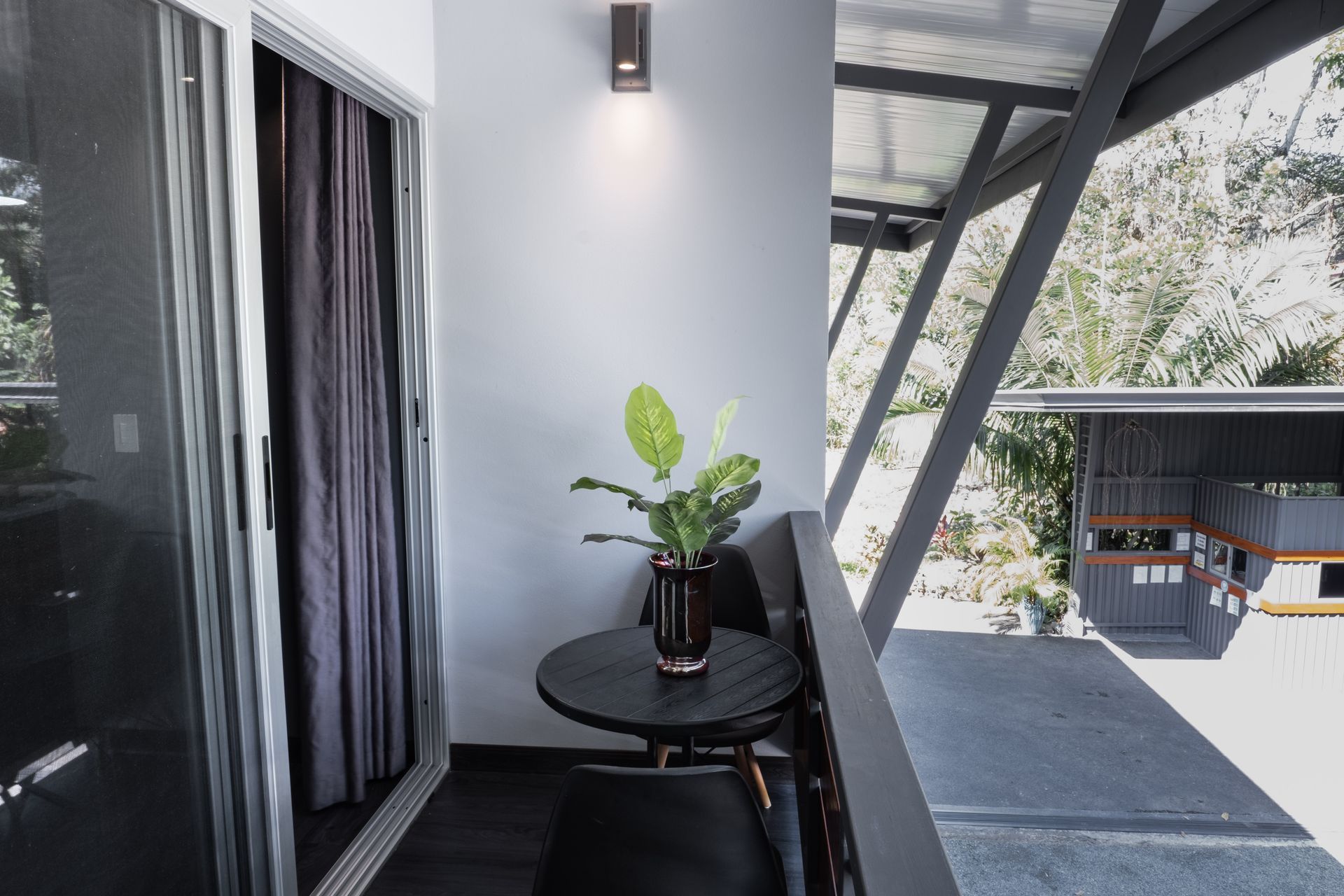 A small balcony features a round black table, two chairs, and a potted plant against a white wall with a wall light.