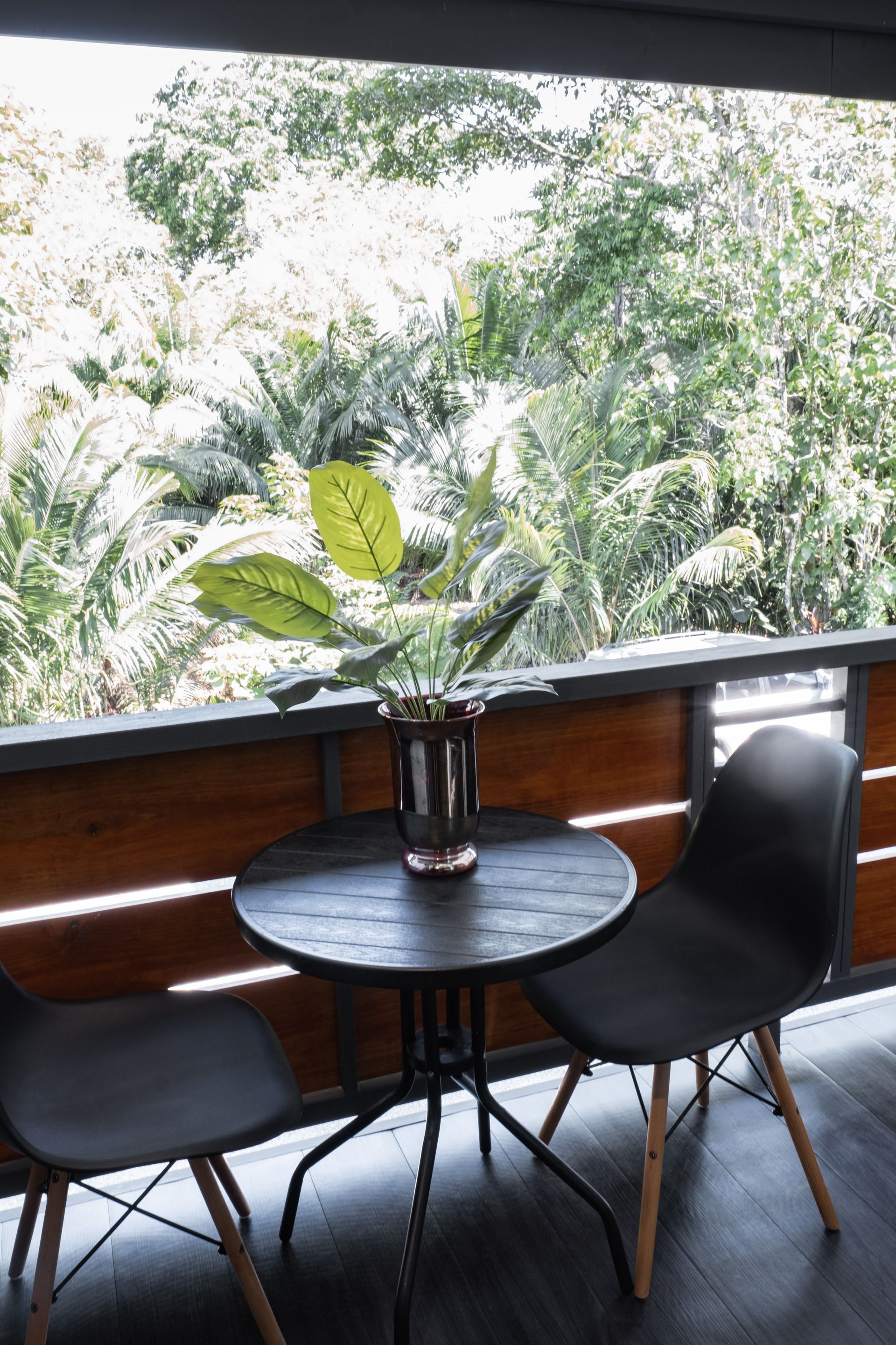 A small, dark round table with two black chairs on a wooden balcony overlooking lush green trees.