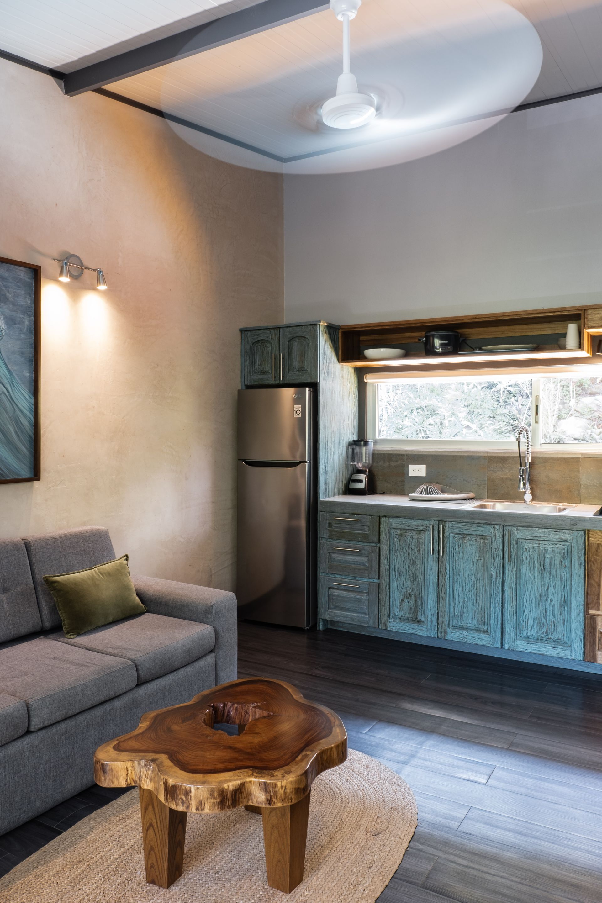 A cozy living room with a gray sofa, a live-edge wooden coffee table on a rug, a refrigerator, and a blue kitchen unit.