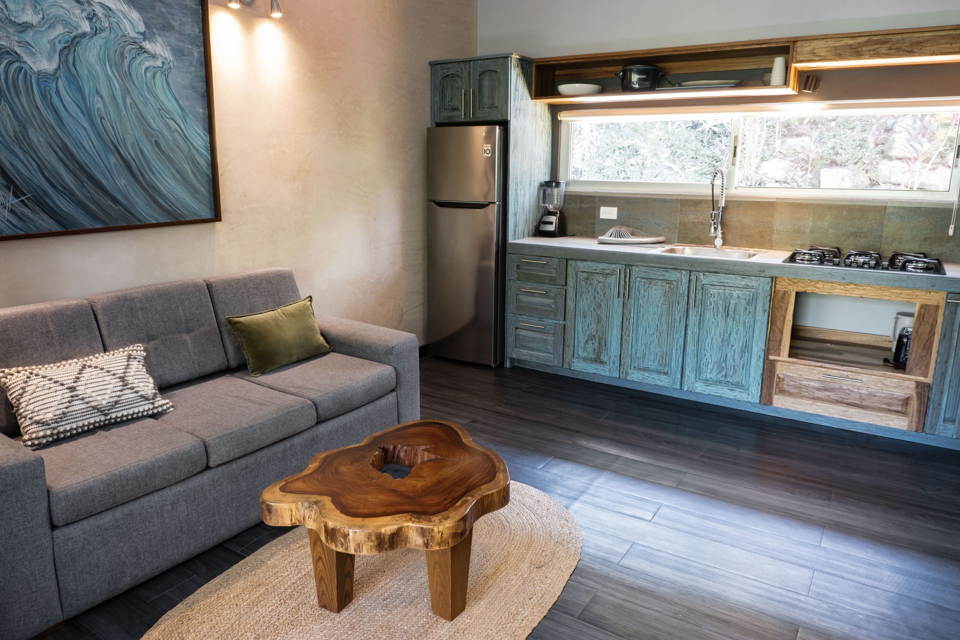A small living area featuring a gray sofa, a live-edge wooden coffee table, a stainless refrigerator, and rustic cabinets.