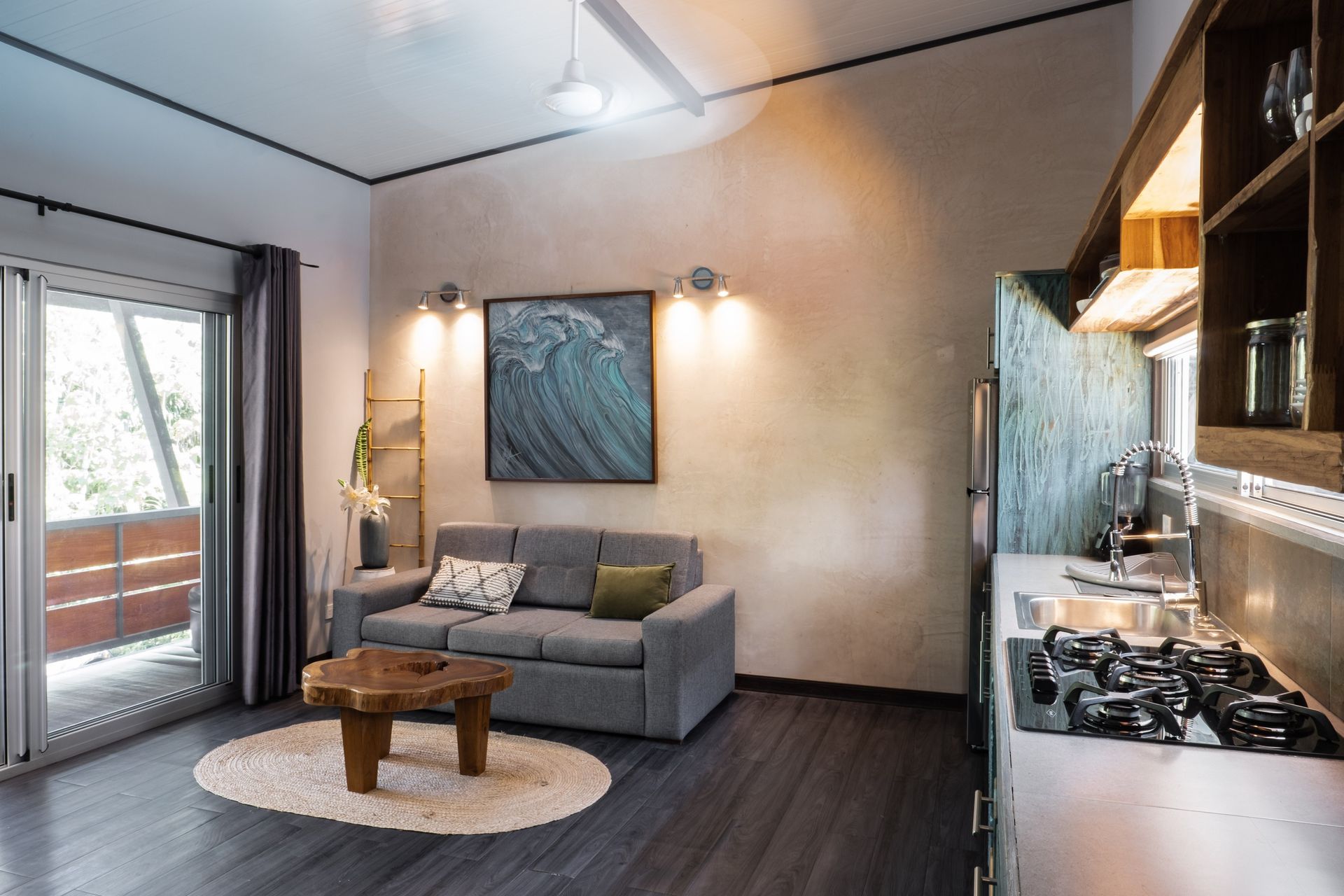 A studio apartment with a grey sofa, live-edge wood coffee table on an oval rug, and a kitchen area with a gas stove.