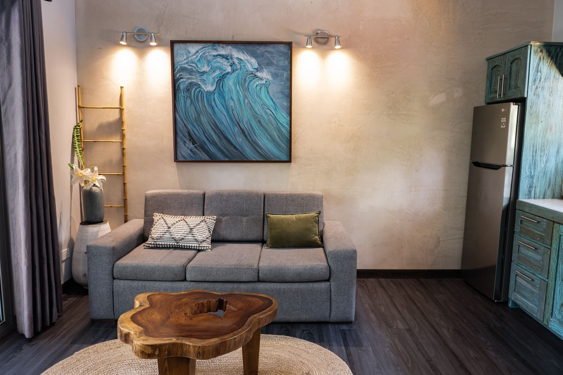 A gray sofa sits in front of a blue wave painting, with a wooden coffee table and a stainless steel refrigerator nearby.
