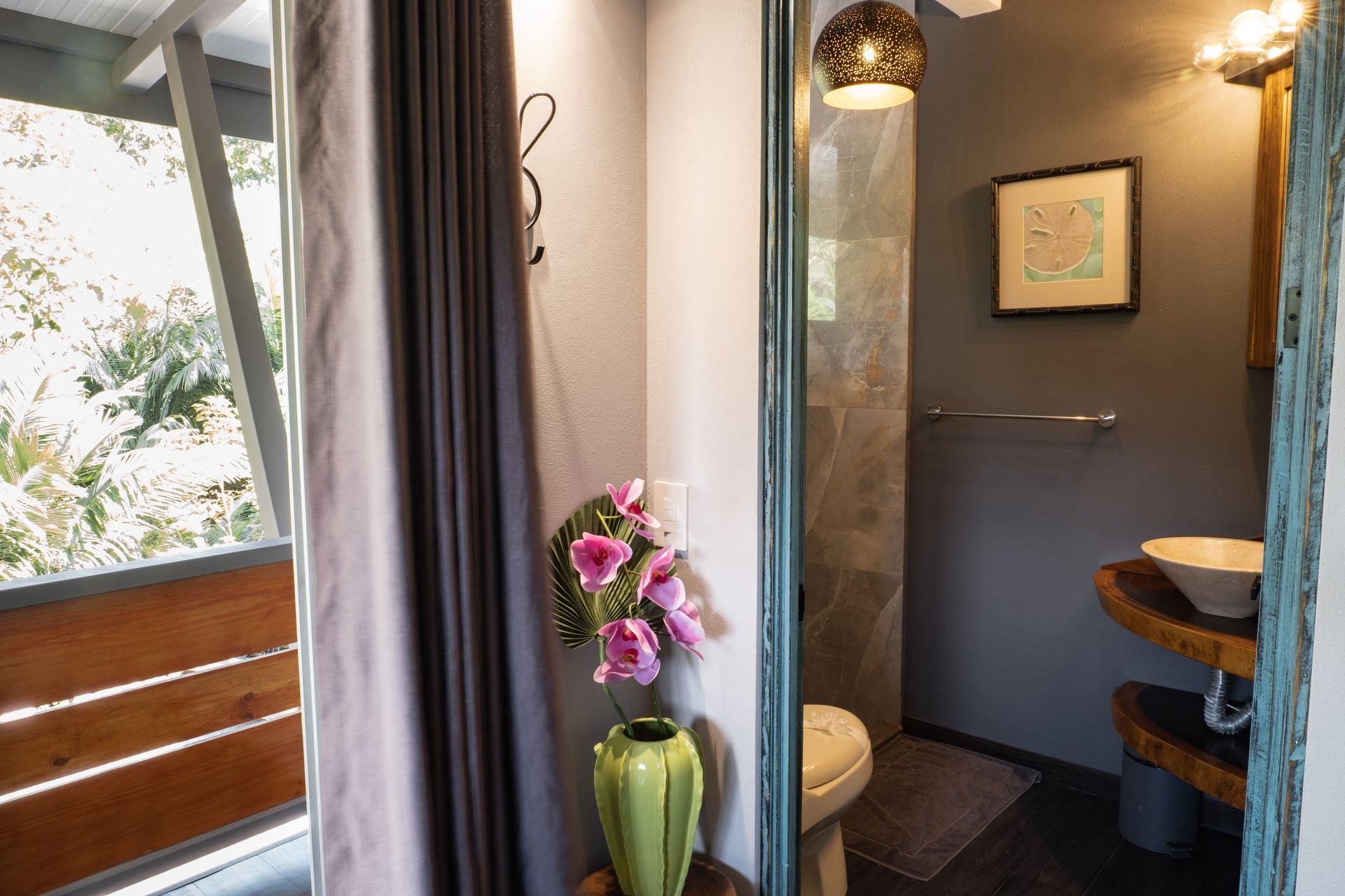 A bathroom with a vessel sink on a live-edge wooden shelf, grey walls, a hanging lantern, and pink flowers in a green vase.
