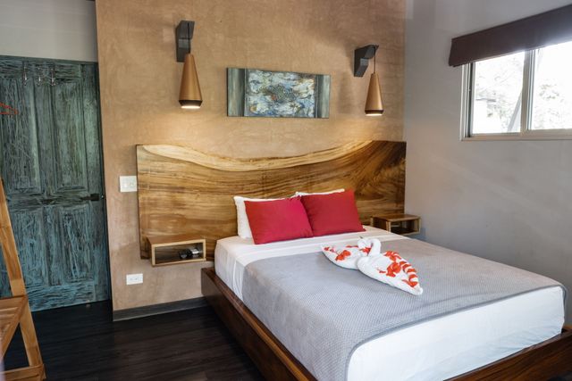 A bedroom with a rustic wood headboard, a white bed with red pillows and towel swans, and two wall lamps on a tan wall.