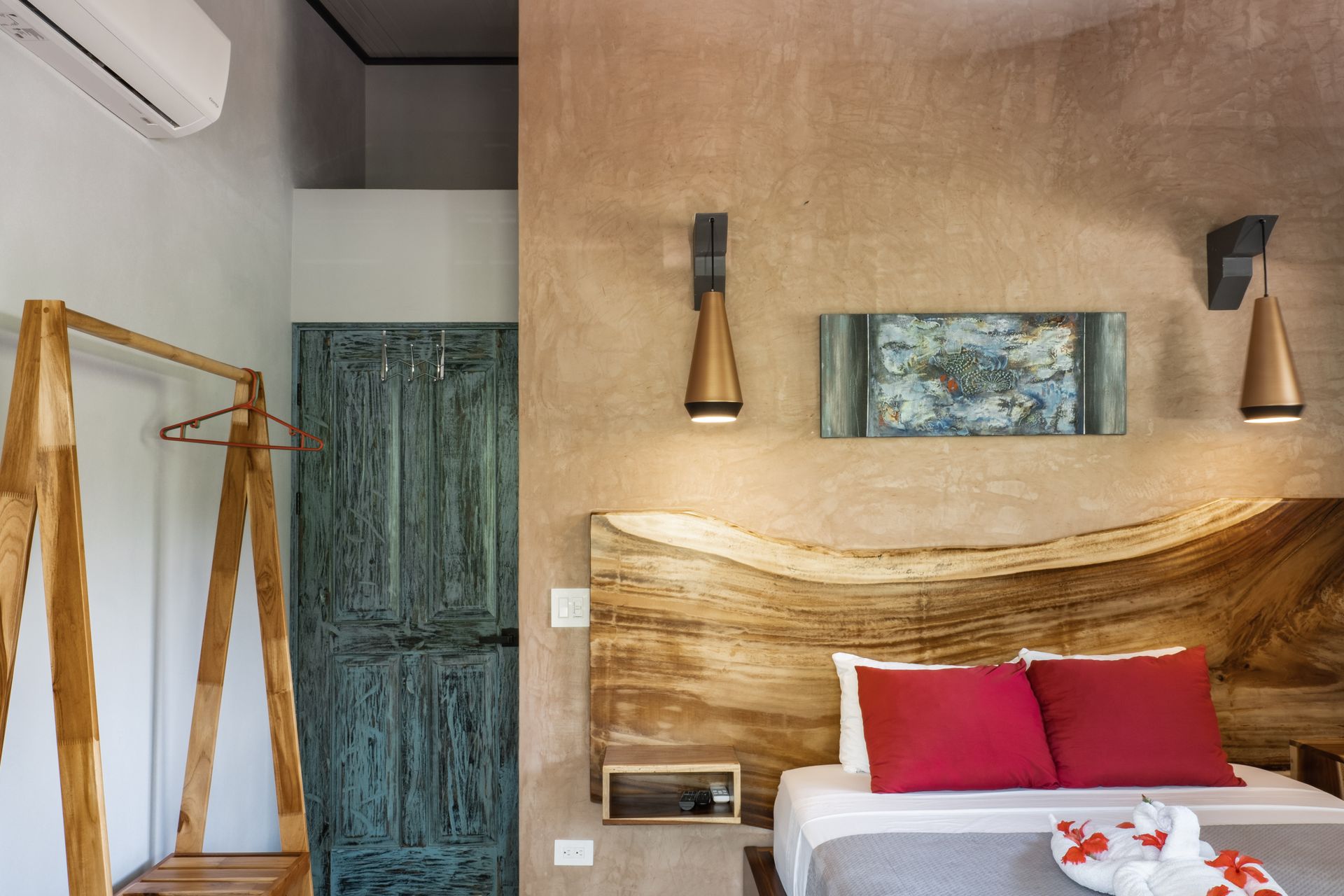 A bedroom with a rustic wooden headboard, two red pillows, warm wall lamps, a green wooden door, and a clothing rack.