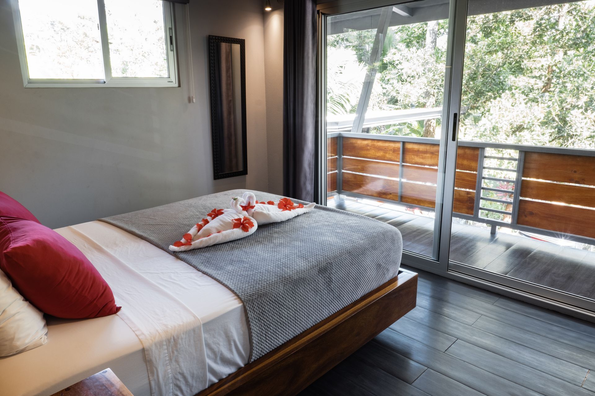 A bedroom with a queen bed featuring towel swans, red pillows, a gray patterned runner, and a sliding door to a balcony.