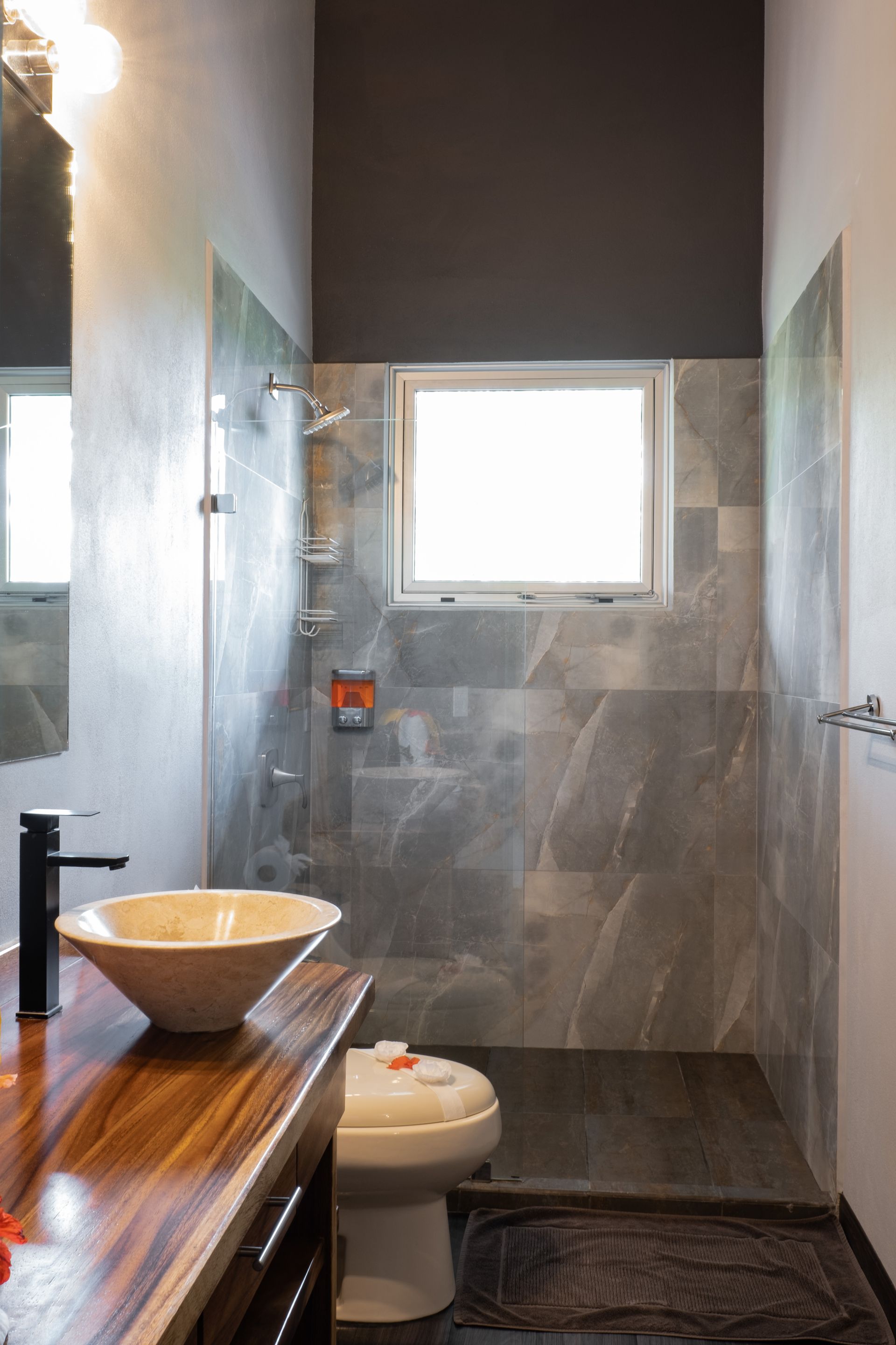 Modern bathroom with stone tile, window shower, white toilet, and wooden vanity with bowl sink
