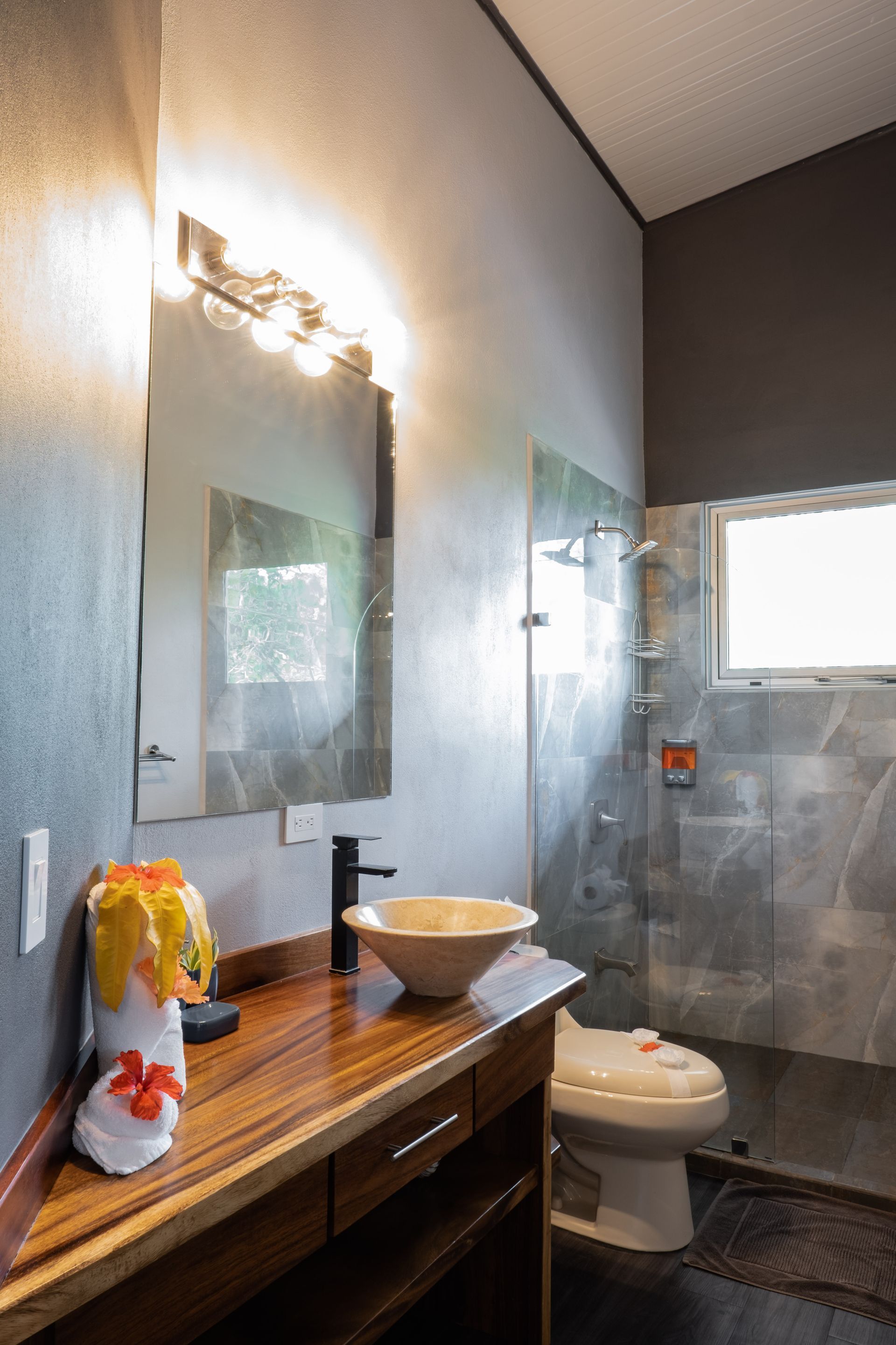 Modern bathroom with wood vanity, white sink, mirror, toilet, and tiled shower area