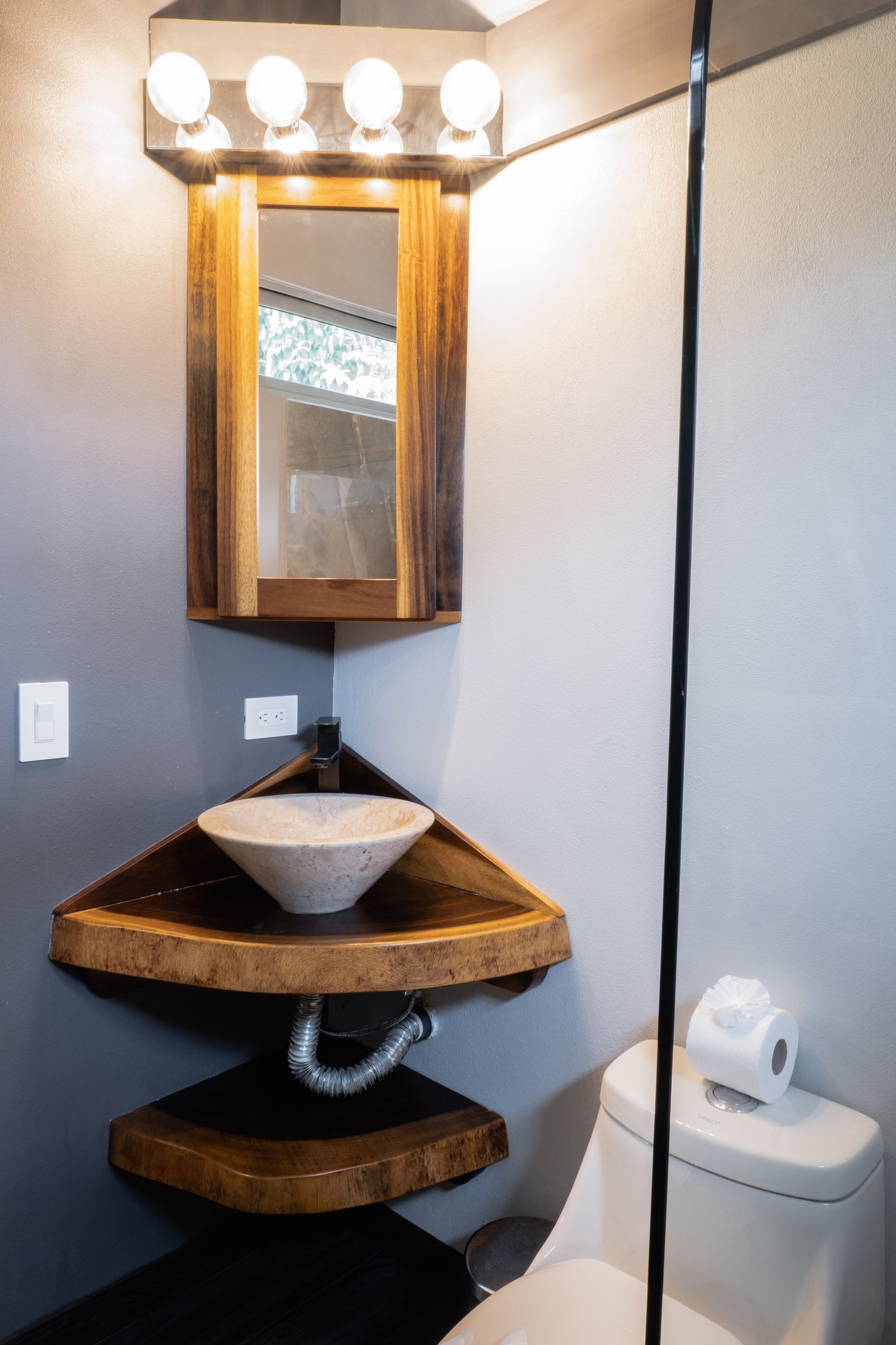 Small bathroom corner with wooden sink shelves, mirror cabinet, vanity lights, and a toilet with toilet paper.