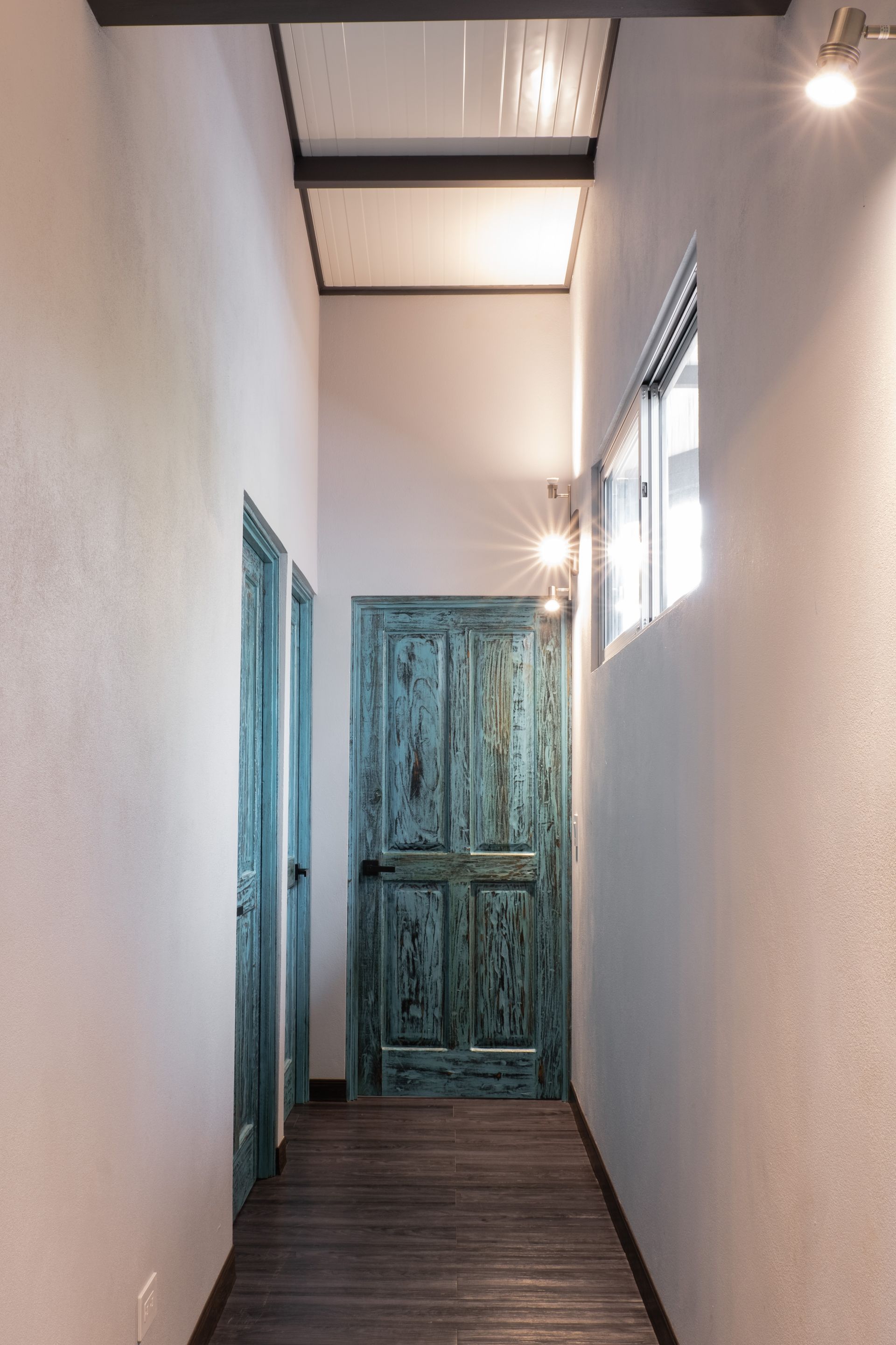 Narrow hallway with teal doors, white walls, wooden floor, and skylight above