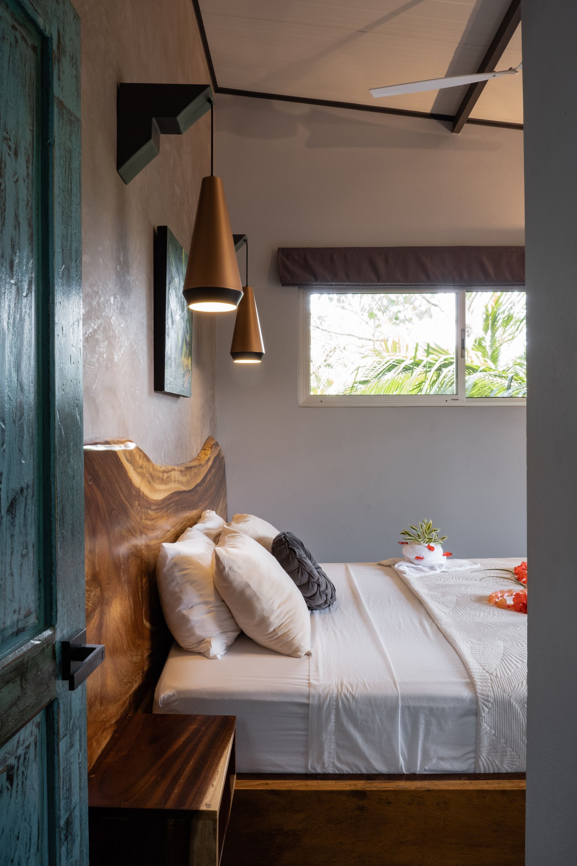 Cozy bedroom with wooden headboard, white bedding, hanging lamps, and a window view of greenery