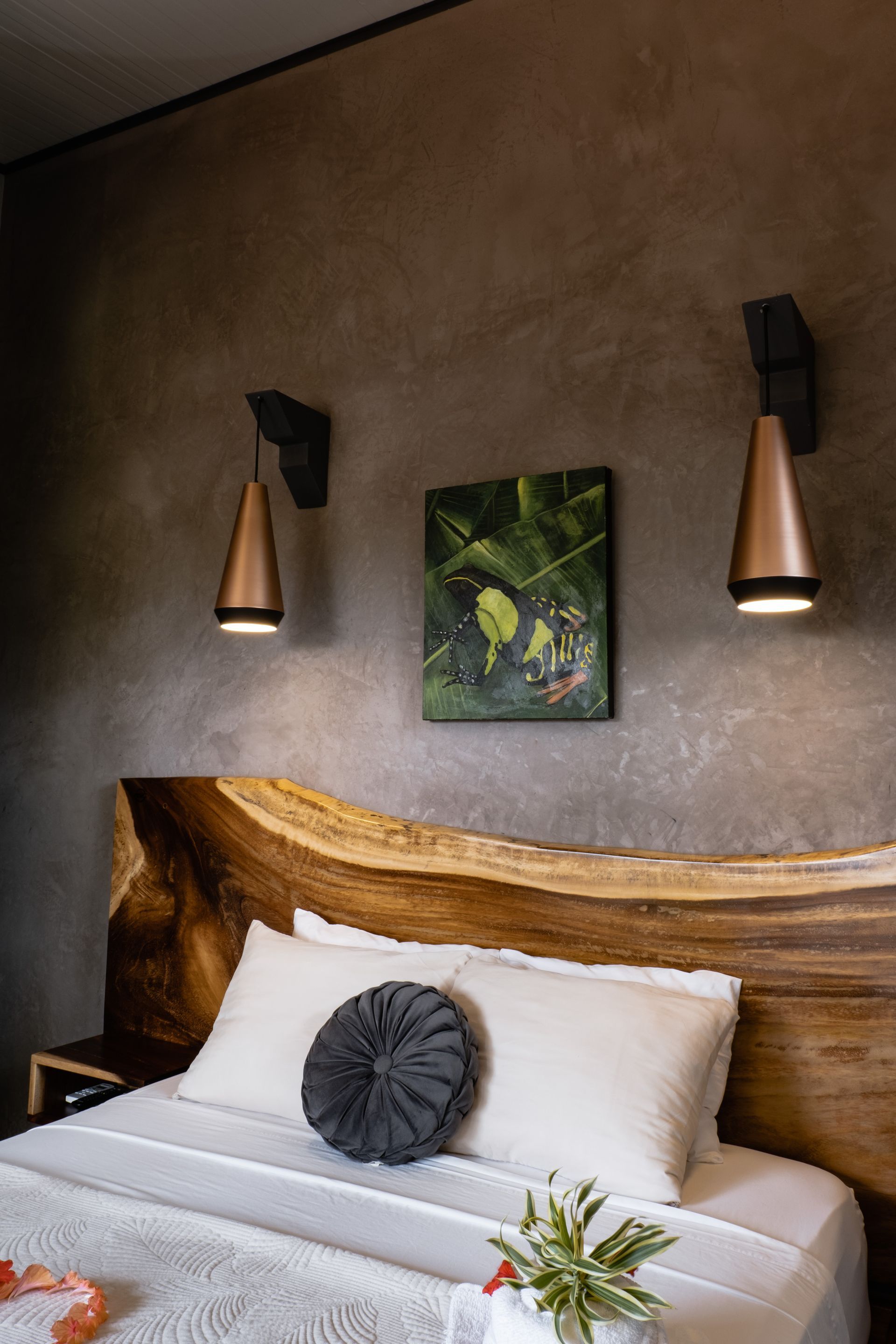 A bedroom features a natural wood slab headboard, two copper pendant lights, and a small botanical painting on a grey wall.