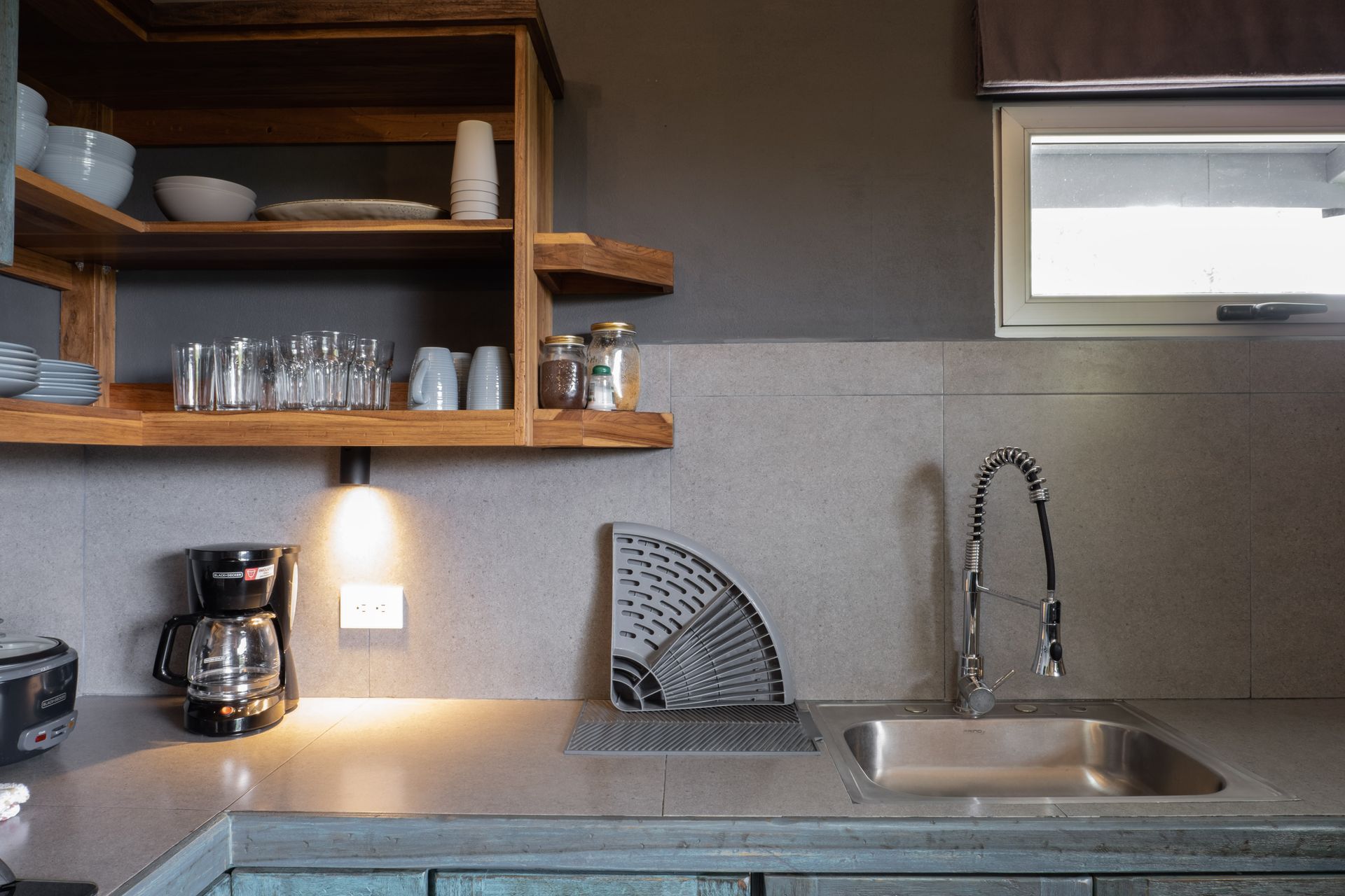 Small kitchen with open shelves, coffee maker, sink, and a window above a beige countertop.