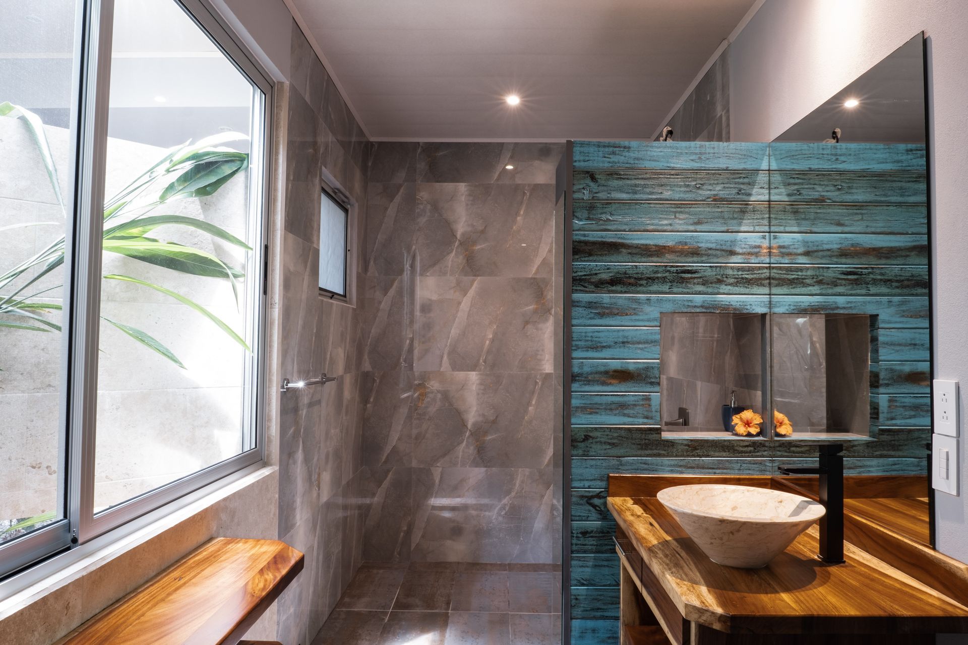 Modern bathroom with gray tile, turquoise accent wall, wood vanity, and a vessel sink by a window.
