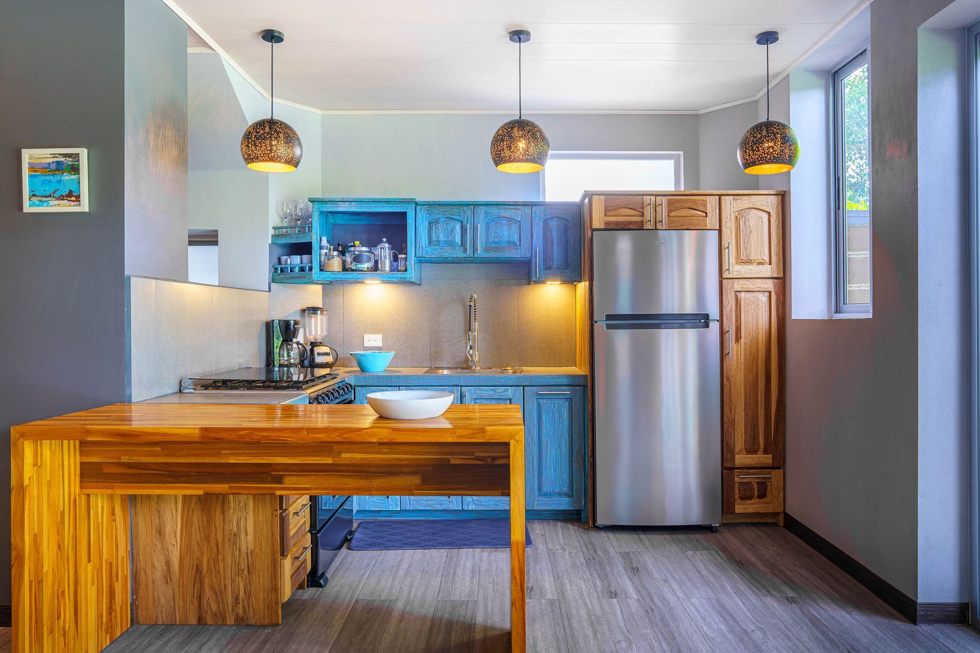 A kitchen with a wood island, blue cabinets, stainless steel refrigerator, and three hanging dome lights.