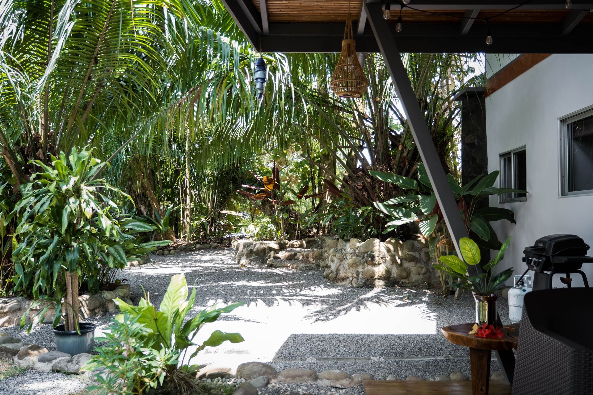 A sunny, gravel patio shaded by lush tropical plants and trees, viewed from under a covered house deck.