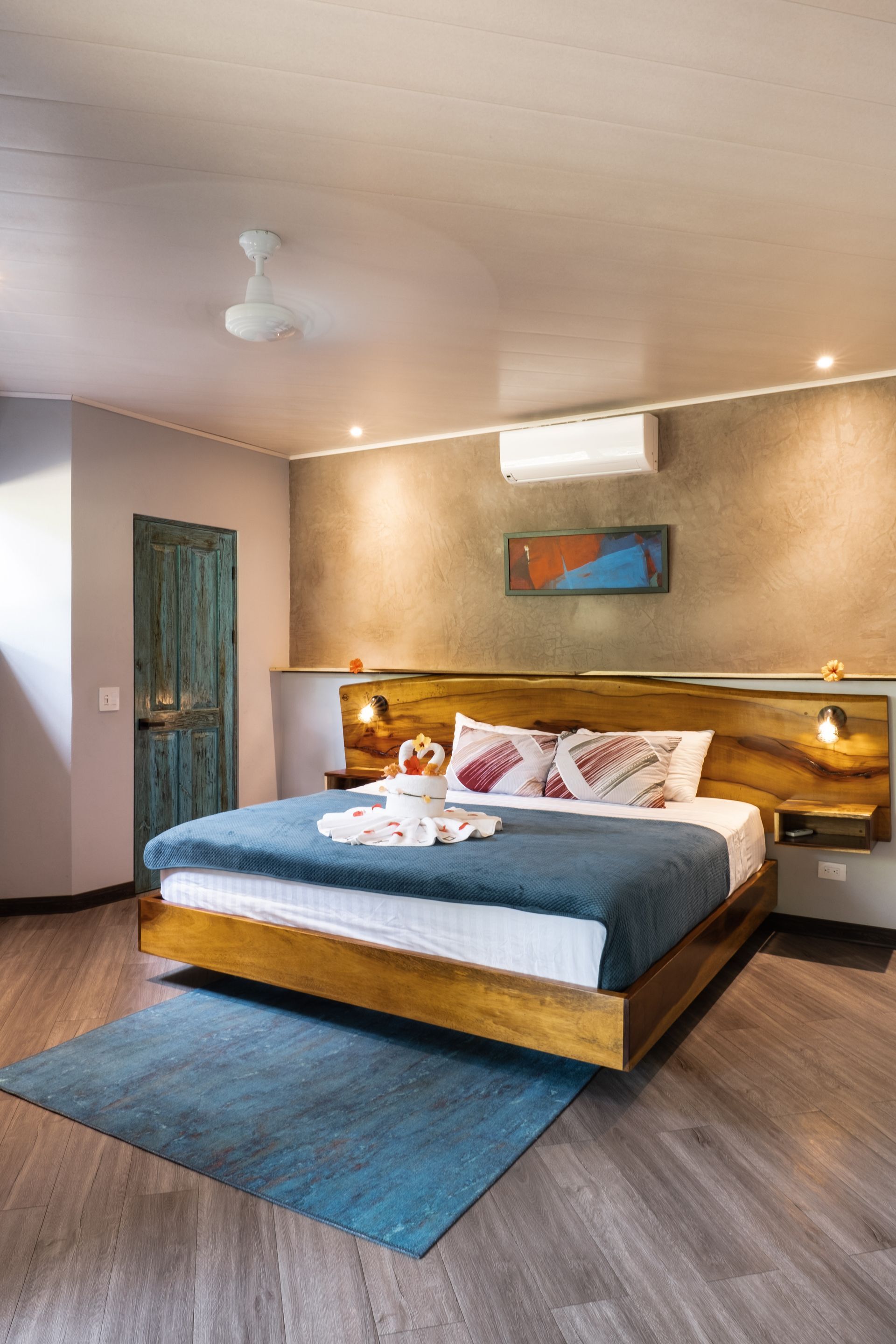 A hotel bedroom featuring a wooden bed with a blue quilt and rug, a textured brown accent wall, and a rustic green door.