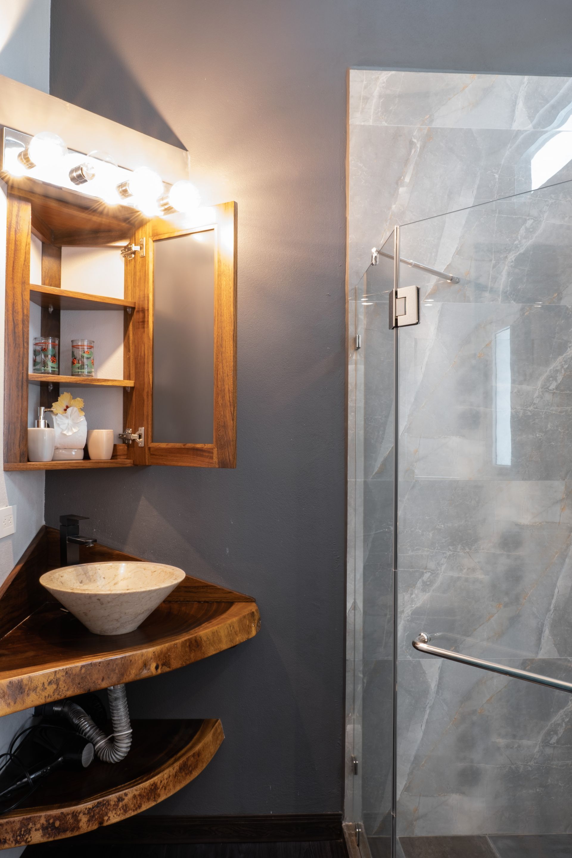 A modern corner bathroom vanity with live-edge wood shelves, a vessel sink, and a mirrored cabinet next to a glass shower.