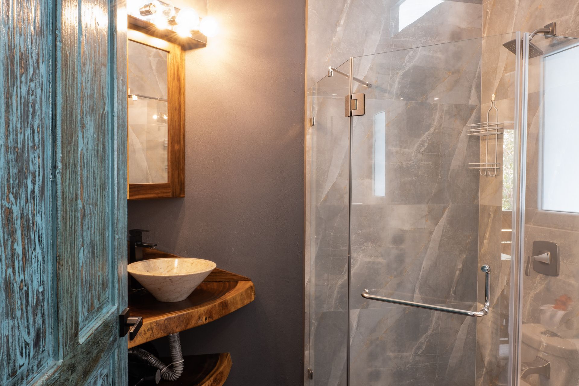A rustic bathroom with a live-edge wooden vanity, white vessel sink, distressed blue door, and glass-enclosed shower.