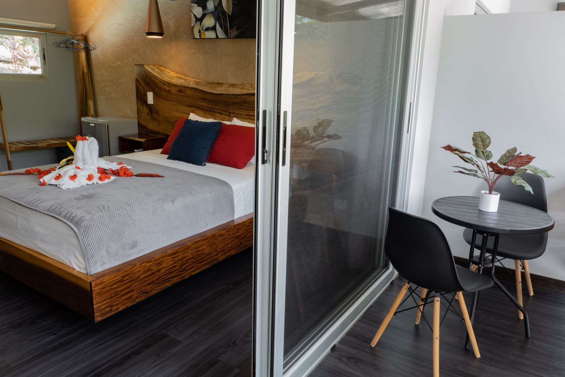 A modern hotel room with a bed featuring a wooden headboard, separated by a glass door from a balcony seating area.
