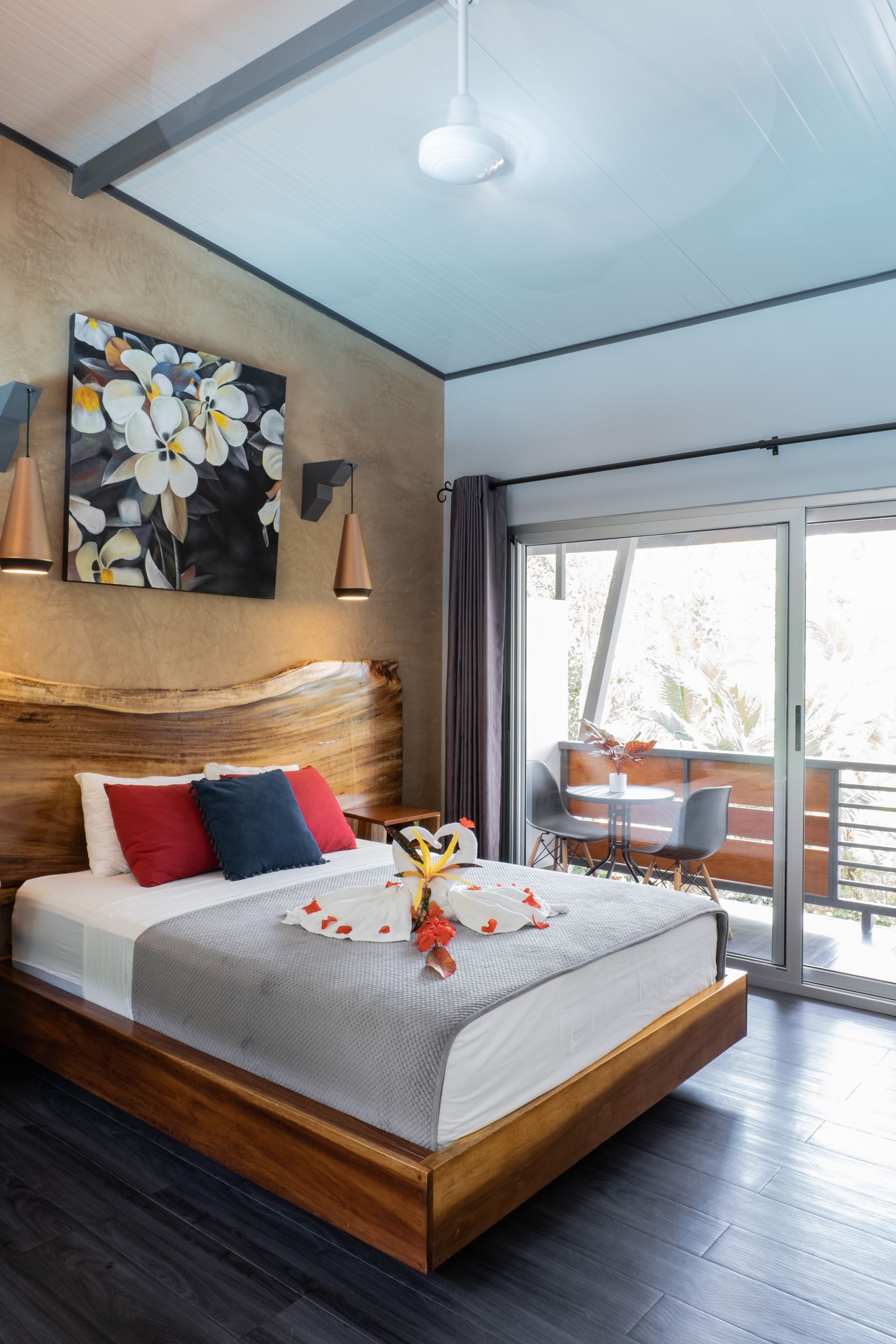 A modern hotel room with a wooden bed frame, red and blue pillows, a flower arrangement, and a balcony view.