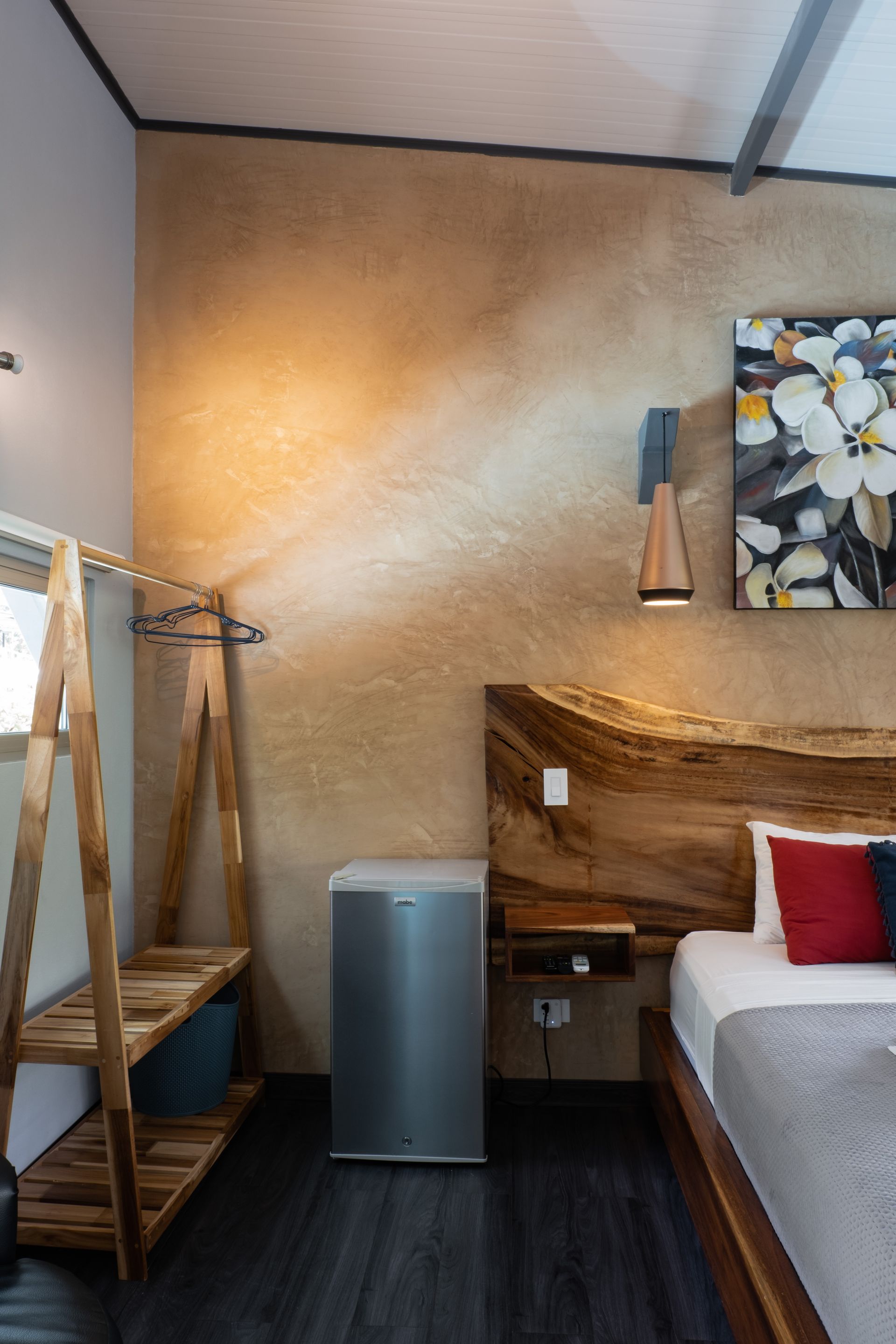A bedroom corner features a wooden A-frame clothes rack, a mini-fridge, and a rustic live-edge headboard with a wall light.
