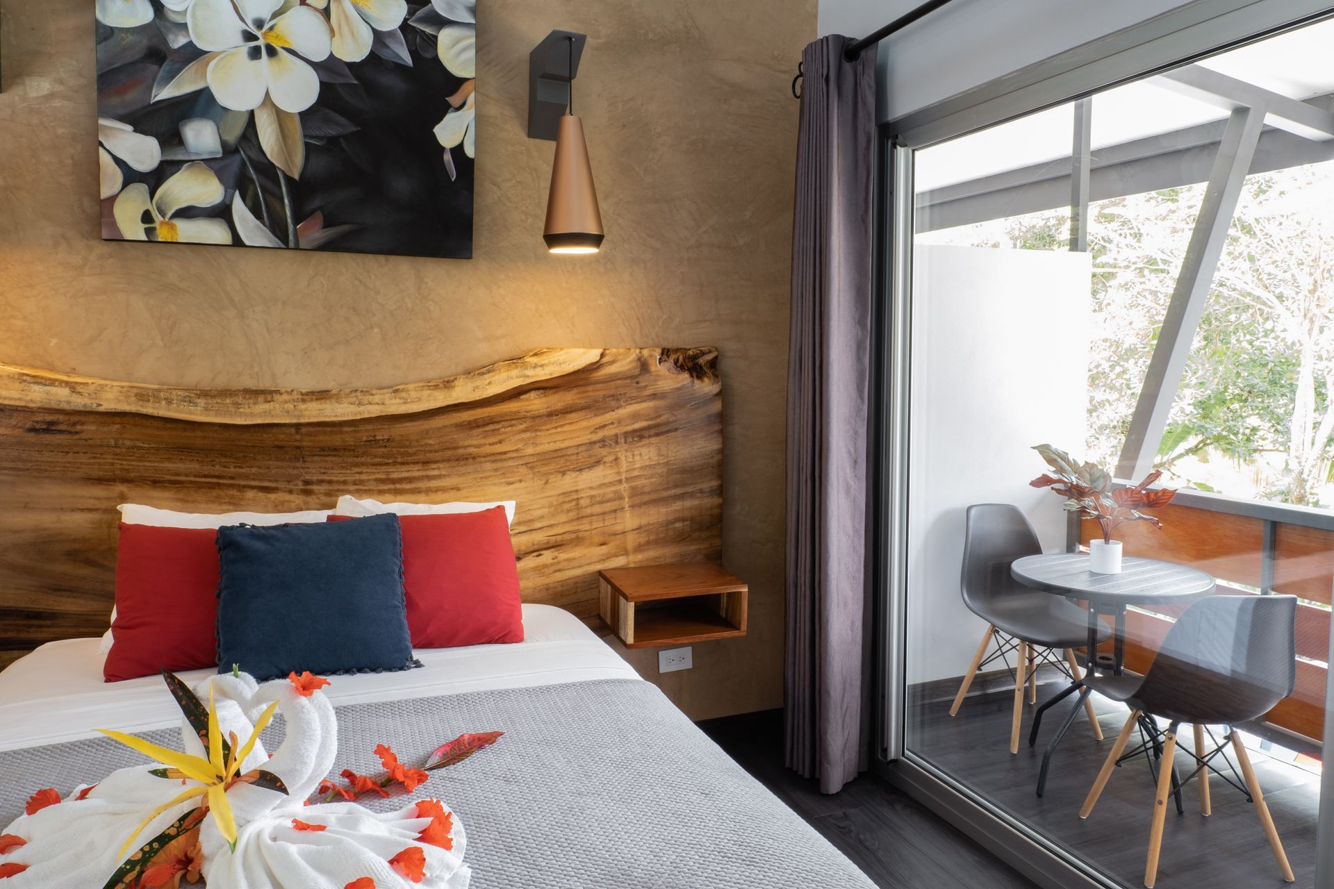 A bedroom with a rustic wood headboard, a white bed with red and blue pillows, and a balcony with a table and chairs.