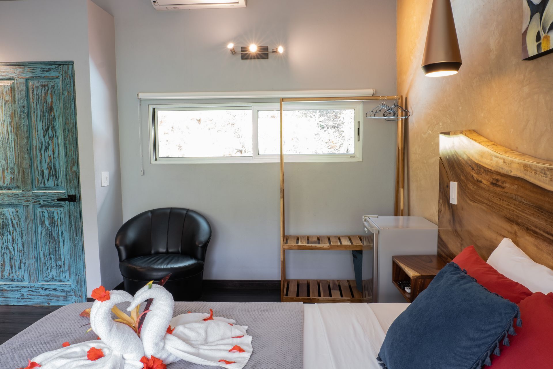 A hotel bedroom with a bed topped with swan-shaped towels, a black armchair, a wooden rack, and a rustic blue door.
