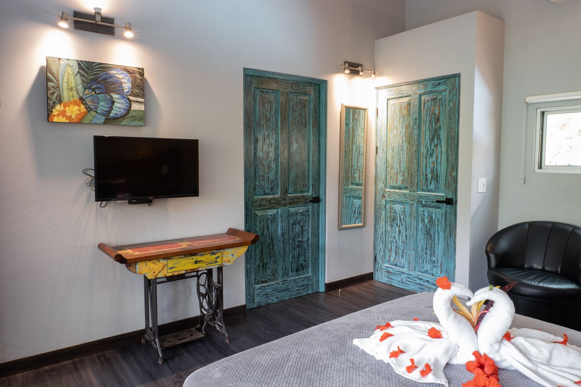 A hotel bedroom featuring a bed with swan-shaped towel art, a wall-mounted TV, and teal rustic doors.