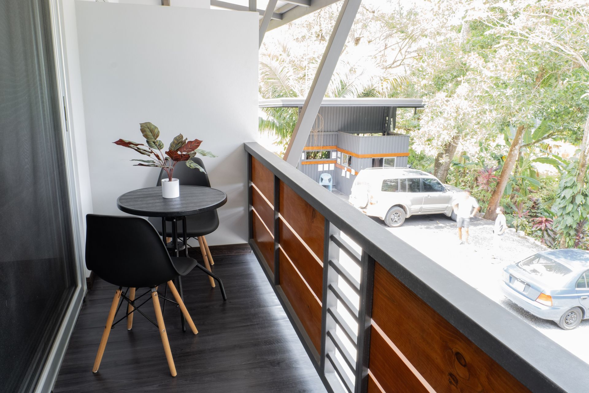 A small balcony with a table, two black chairs, and a potted plant, overlooking a parking area with trees and vehicles.