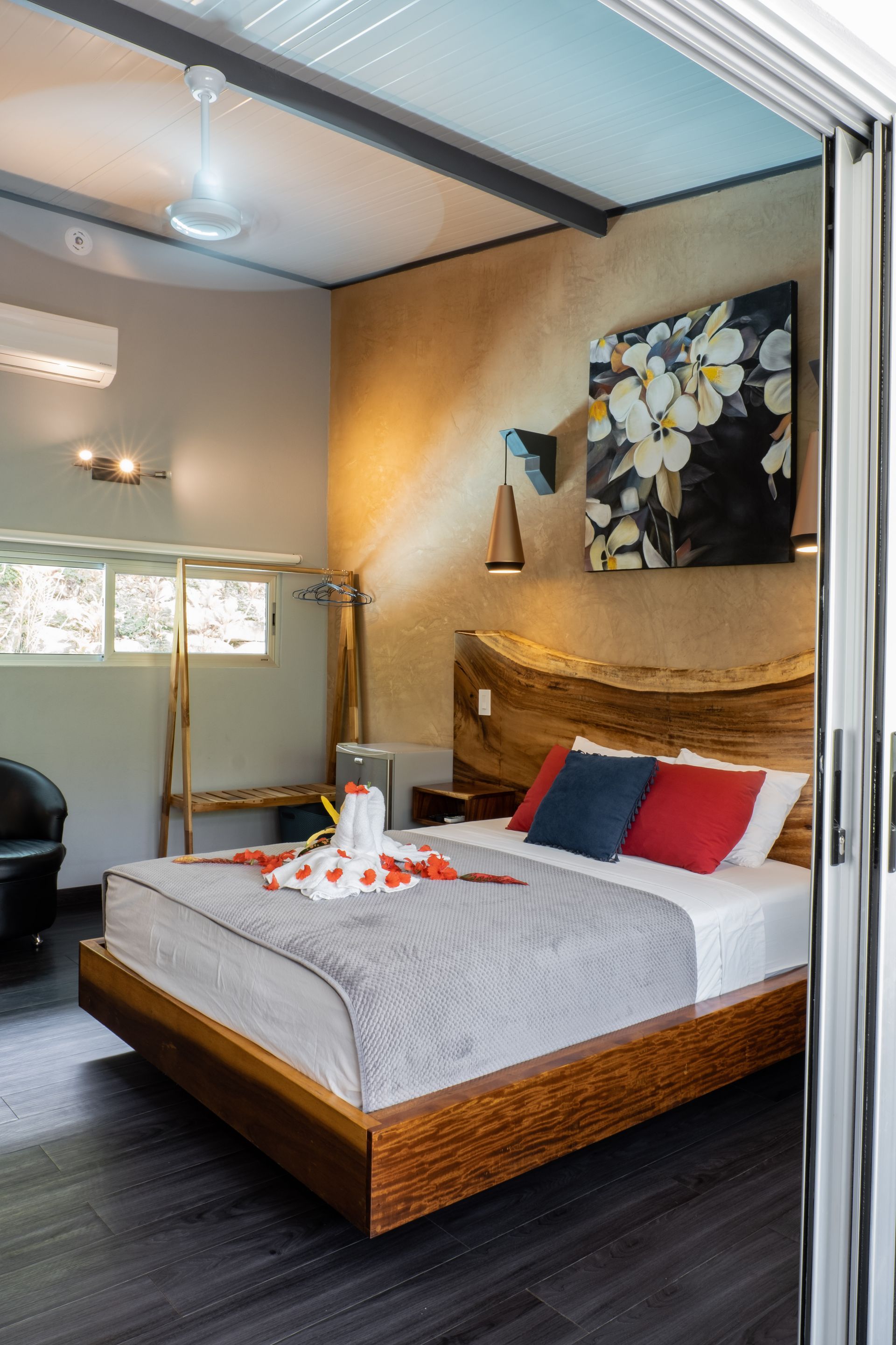 A modern bedroom with a wooden live-edge headboard, a white bed with colorful pillows, and a large floral wall canvas.