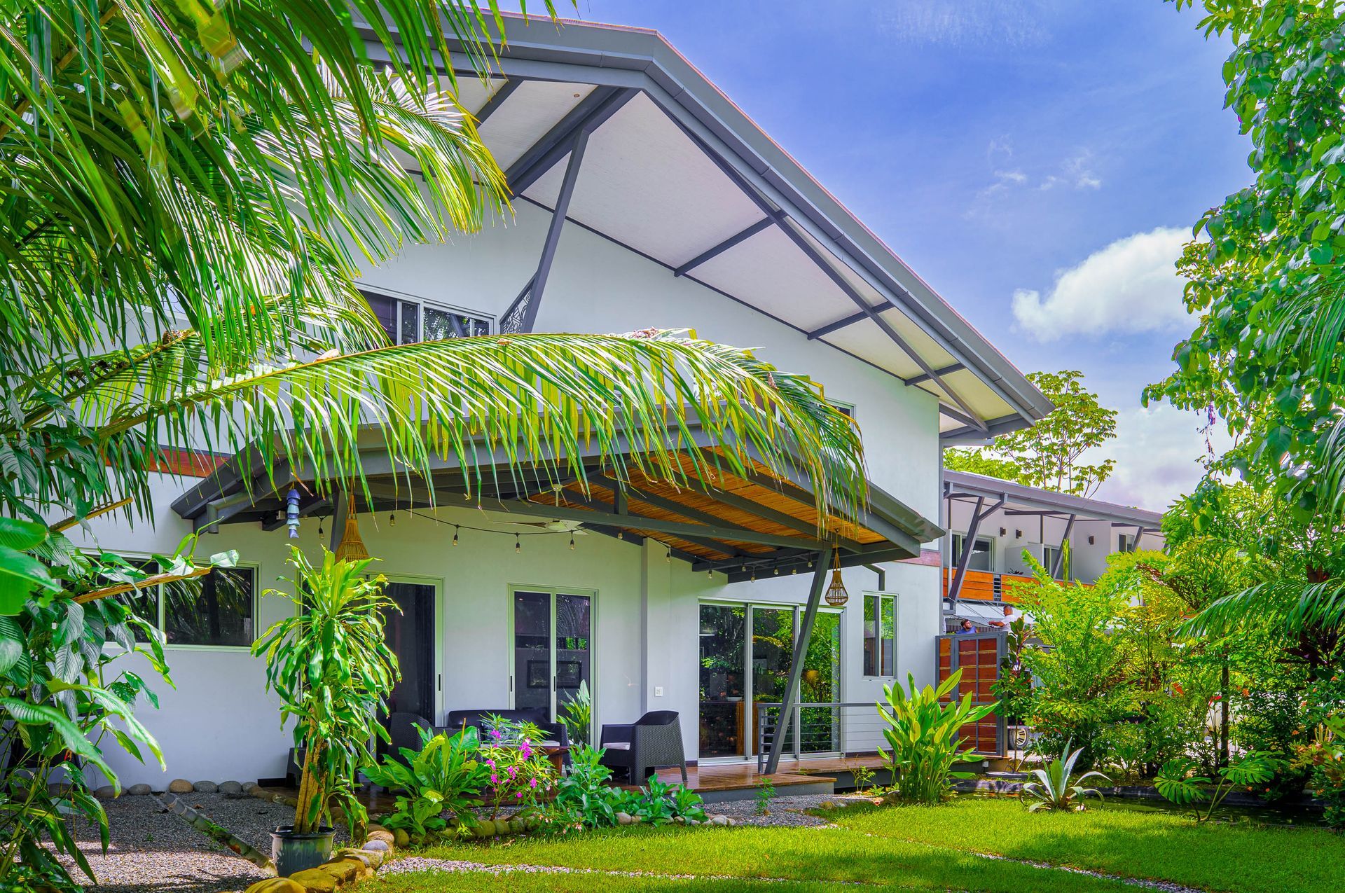 A white, multi-level house with a patio roof, surrounded by lush green foliage and trees under a blue sky.