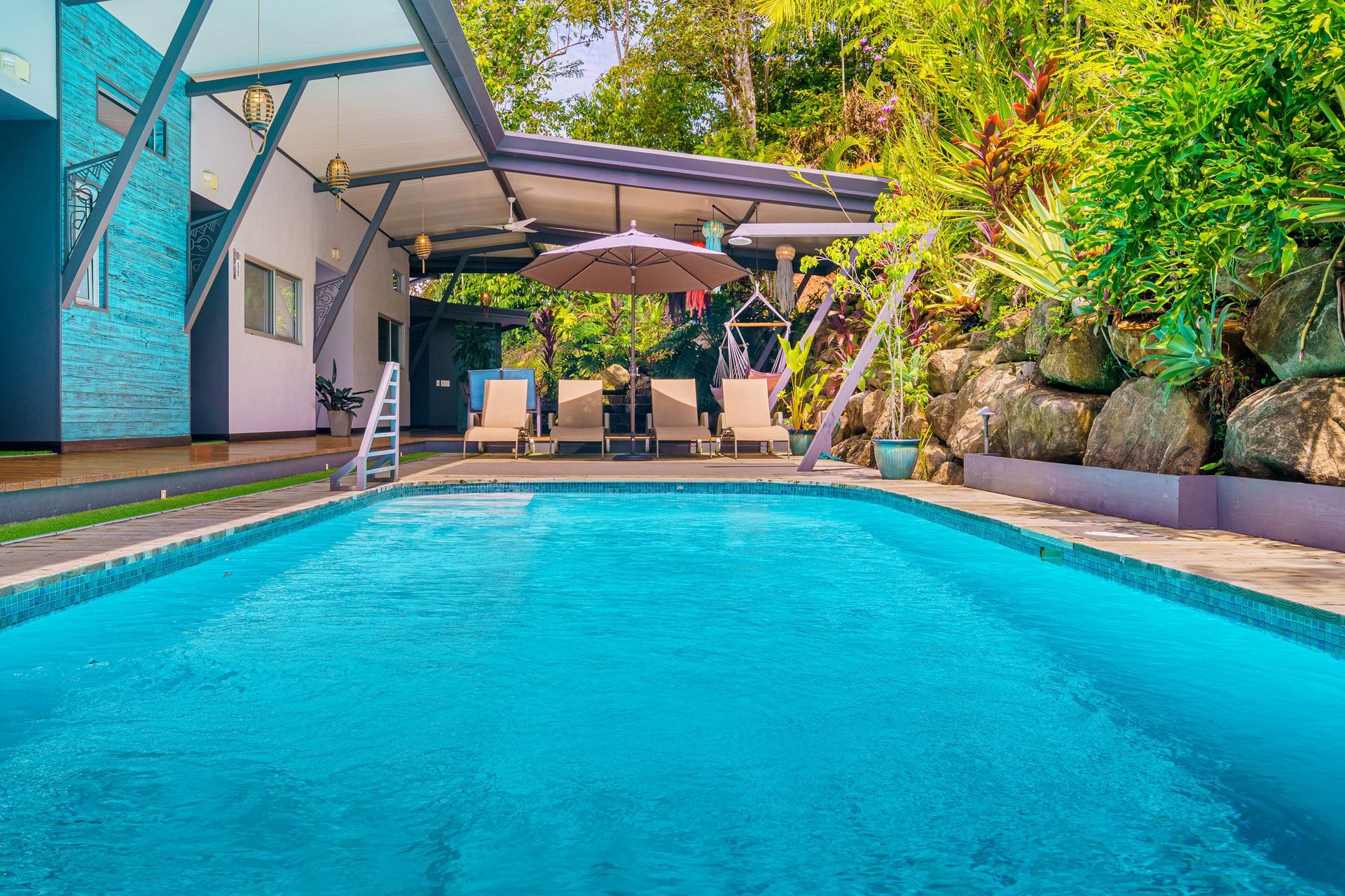 A bright blue rectangular swimming pool in a tropical setting with lounge chairs under a patio roof.