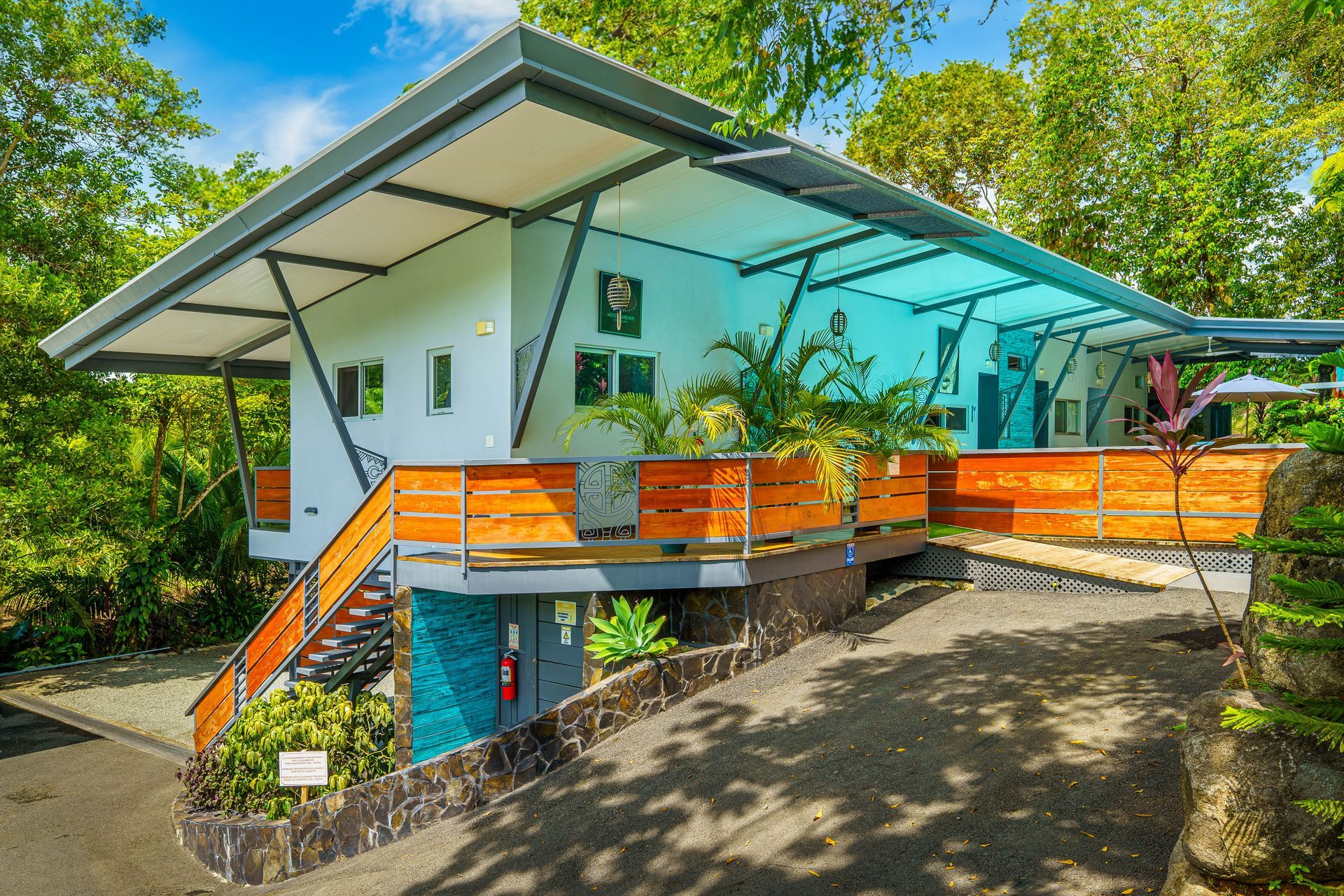 Modern house with a bright blue metal roof, white walls, and wood-paneled balcony, surrounded by lush green trees.