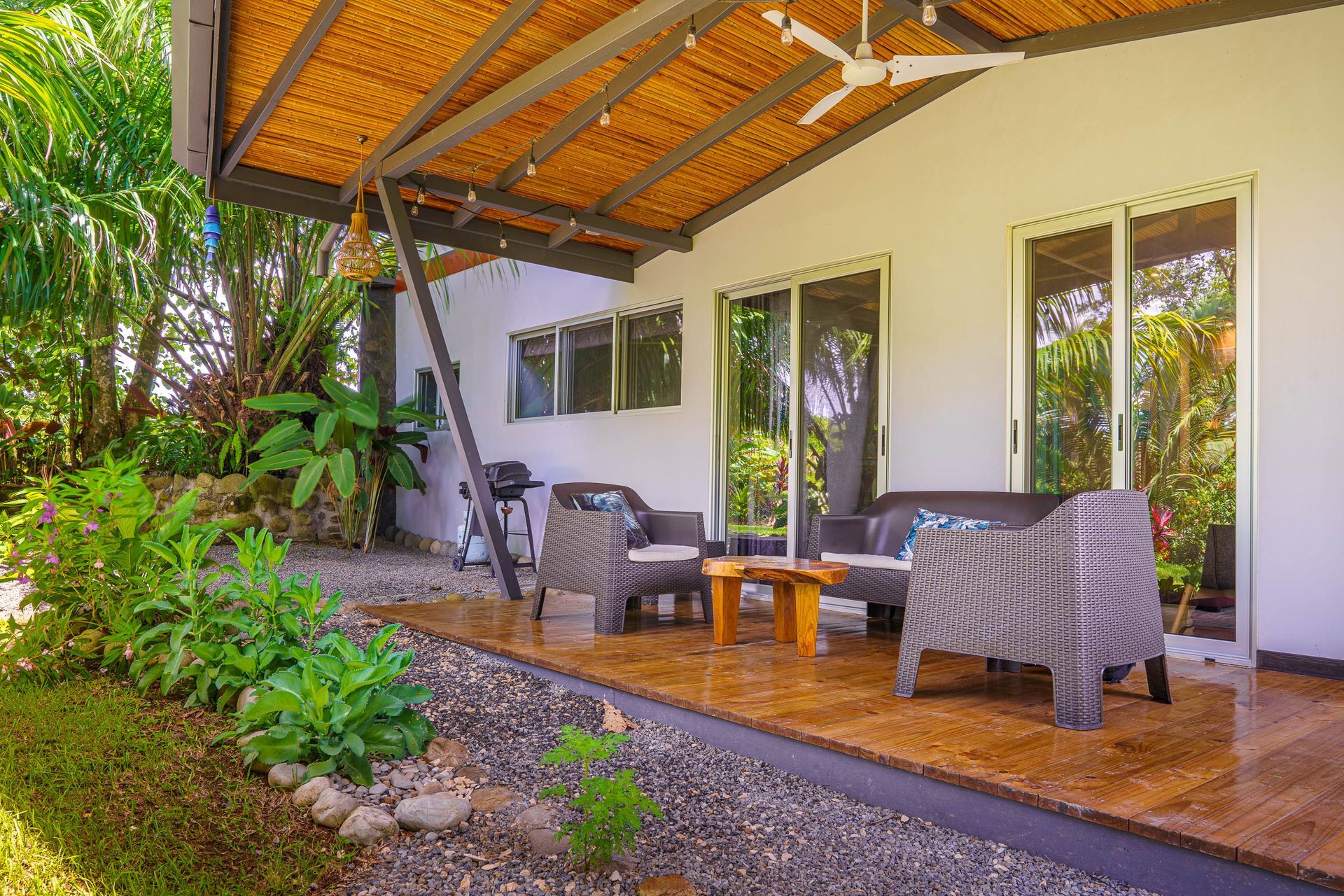 Covered patio with wicker chairs, wooden table, and lush garden beside a white house