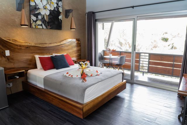 A modern bedroom featuring a wooden headboard, a white bed with red and blue pillows, and a balcony with outdoor seating.