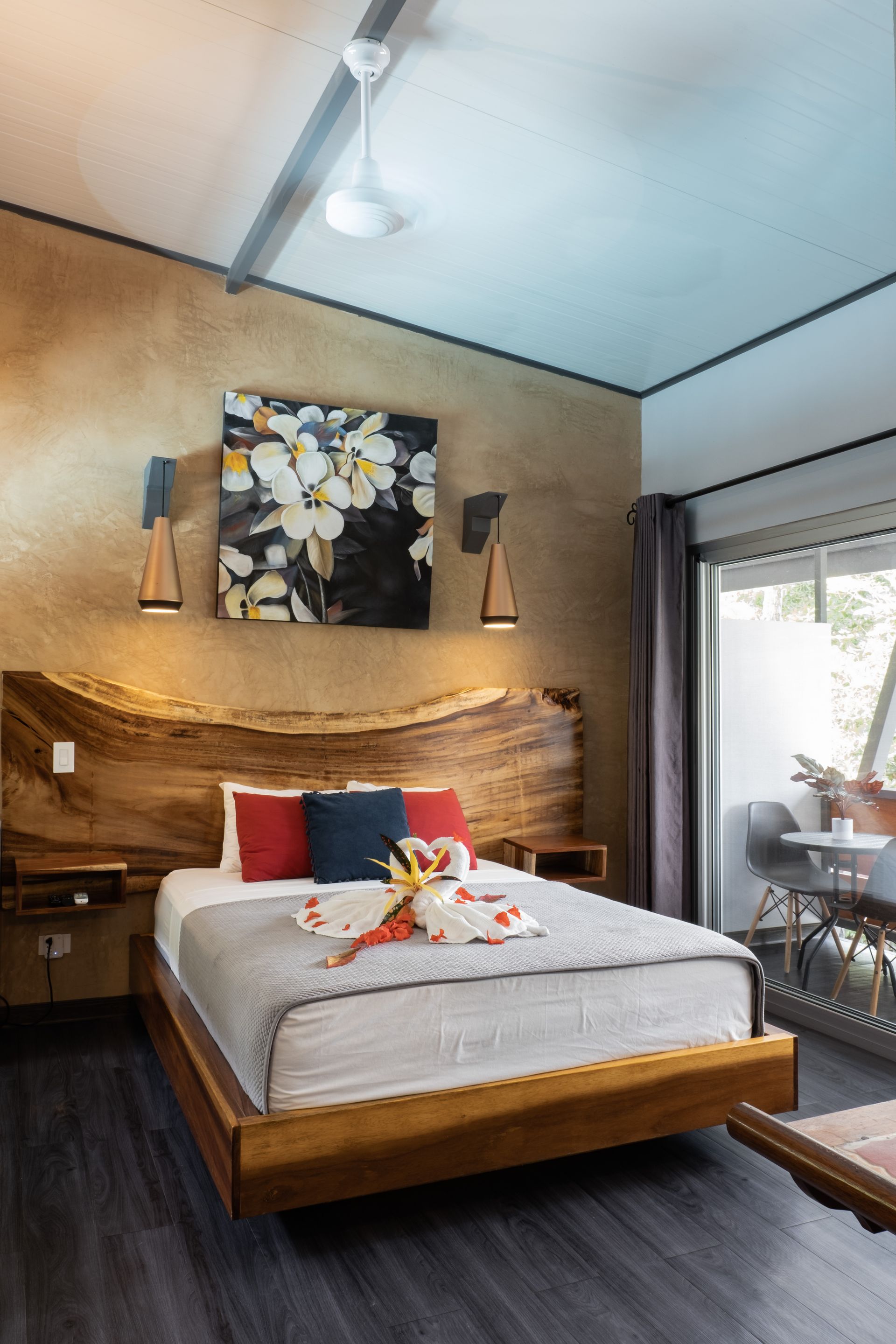 A modern hotel bedroom with a live-edge wood headboard, floral art, warm lighting, and a view of an outdoor patio.