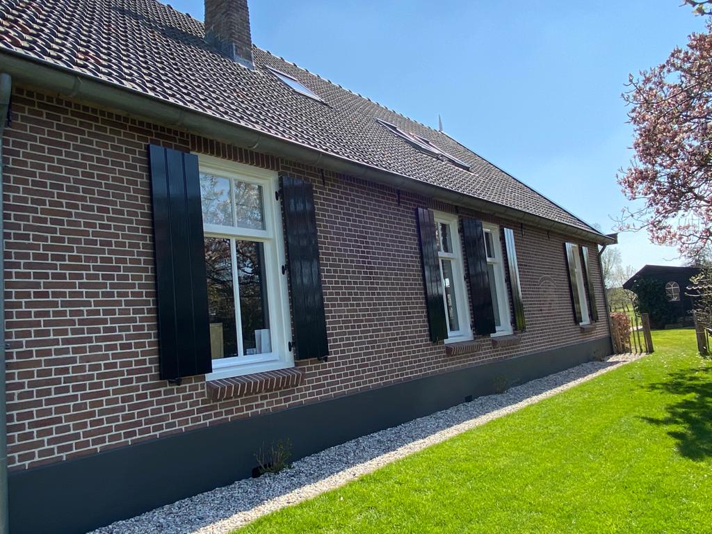 De buitenkant van een bakstenen huis met geschilderde luiken, witte ramen en een groen gazon onder een blauwe hemel.