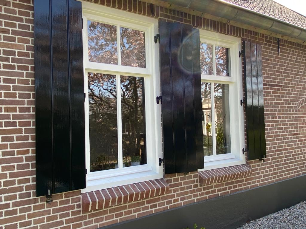 Een bakstenen huismuur met witte kozijnen en geschilderde luiken.