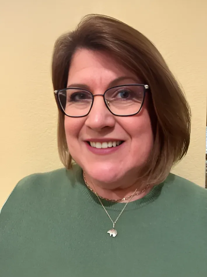 Woman with brown hair and glasses, smiling, wearing a green shirt and gold necklace.