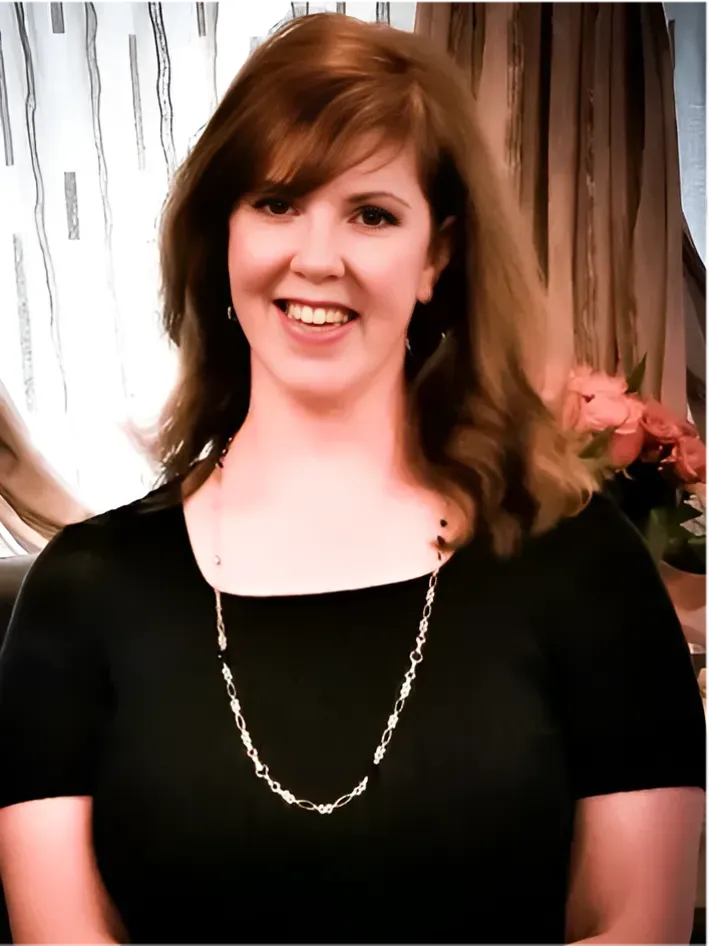 Woman with red hair, wearing a black top and silver necklace, smiling. Background includes flowers and curtains.