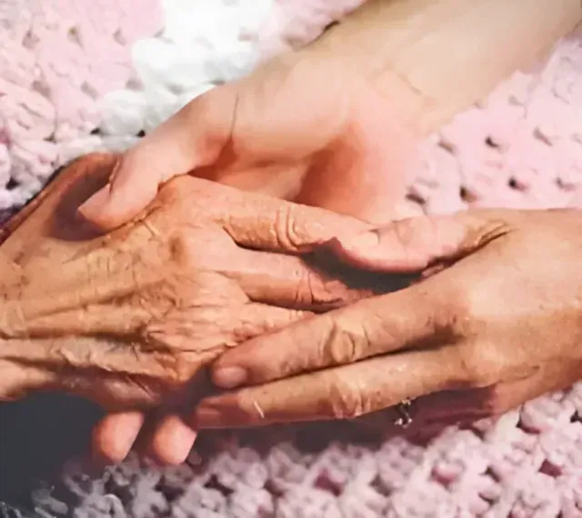 Two hands, one aged and wrinkled, held by a younger hand, resting on a pink, crocheted blanket.