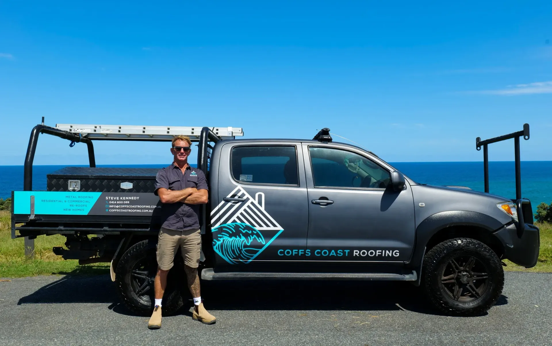 Man standing beside a grey work truck with logo near ocean.