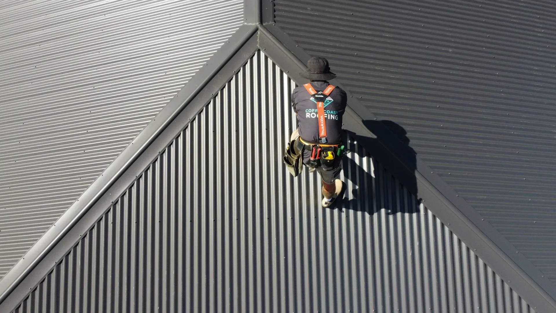 Roofer on a gray corrugated metal roof, wearing safety harness.