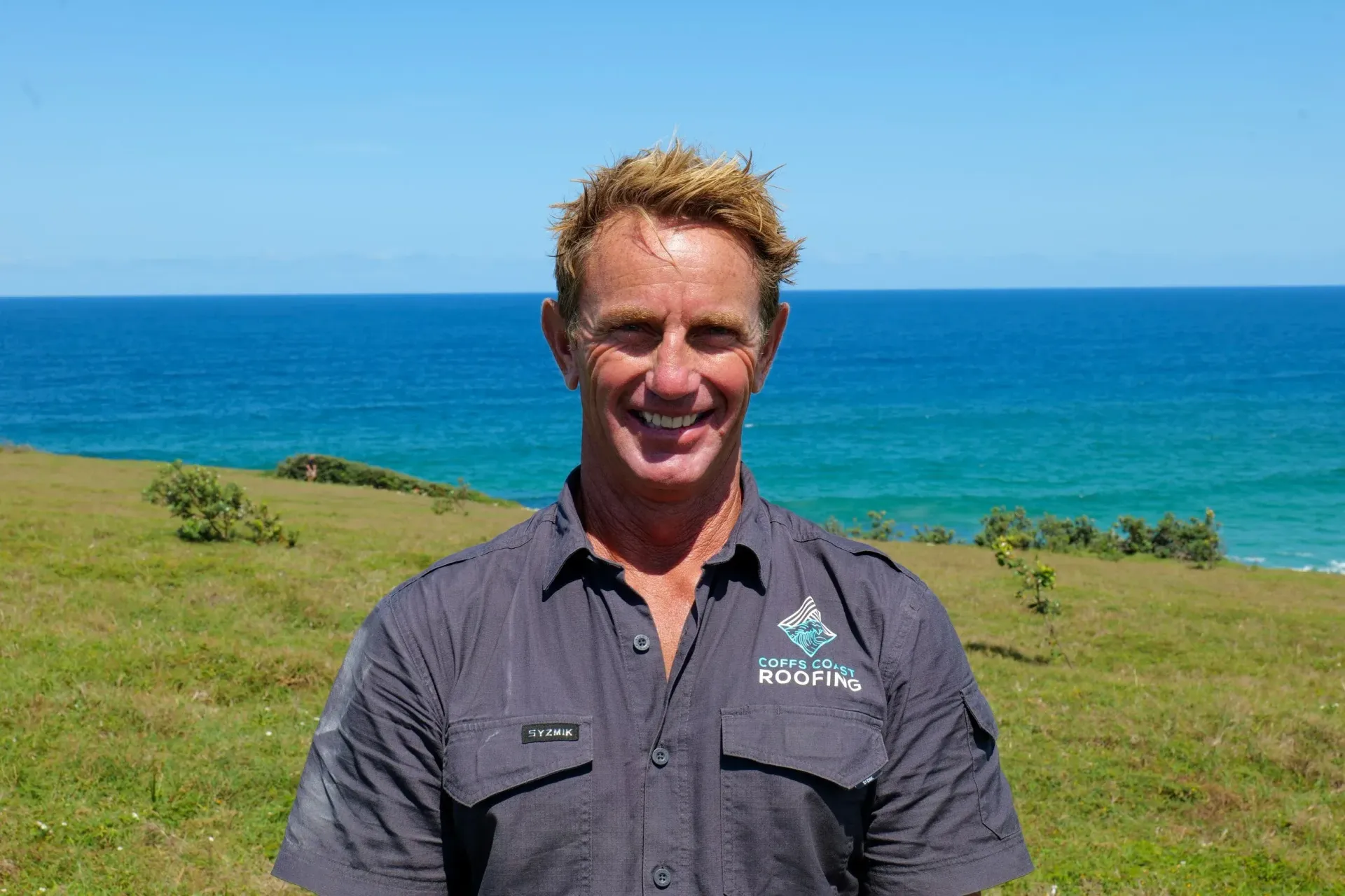 Man in grey shirt smiles in front of ocean and green hillside.