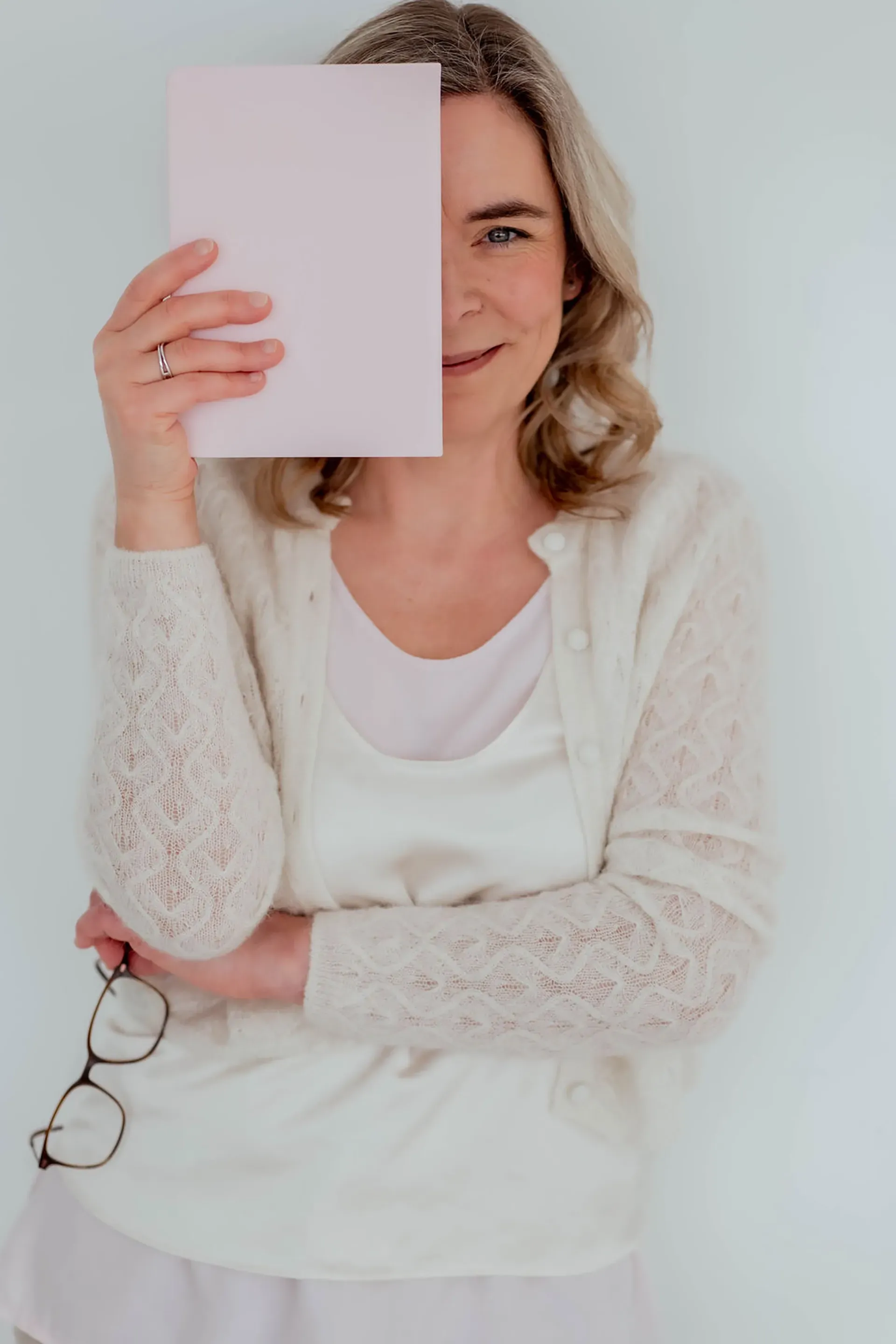 Frau hält ein leeres Blatt Papier vor einen Teil ihres Gesichts; sie steht vor einem hellen Hintergrund.